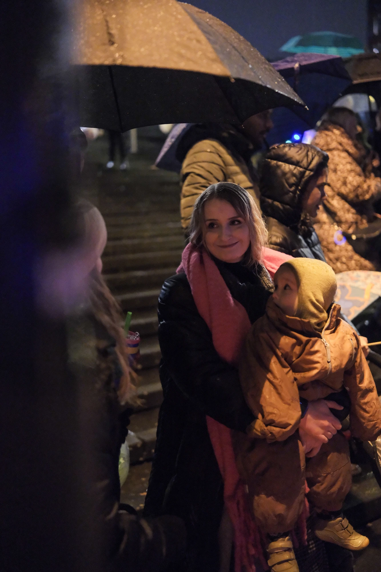 Frau mit rosa Schal hält Kind in gelber Kapuzenjacke, mehrere Personen mit Regenschirmen im Hintergrund bei Nacht.