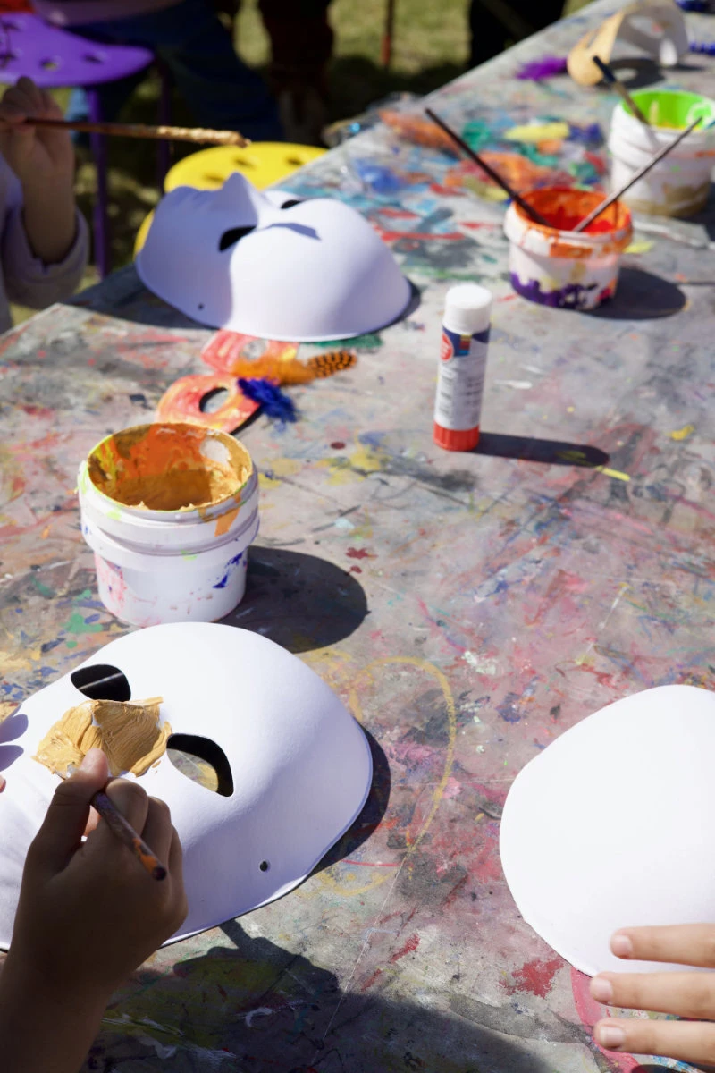 Kinder bemalen weiße Masken auf einem mit Farbflecken bedeckten Tisch mit Pinseln und Farbe.