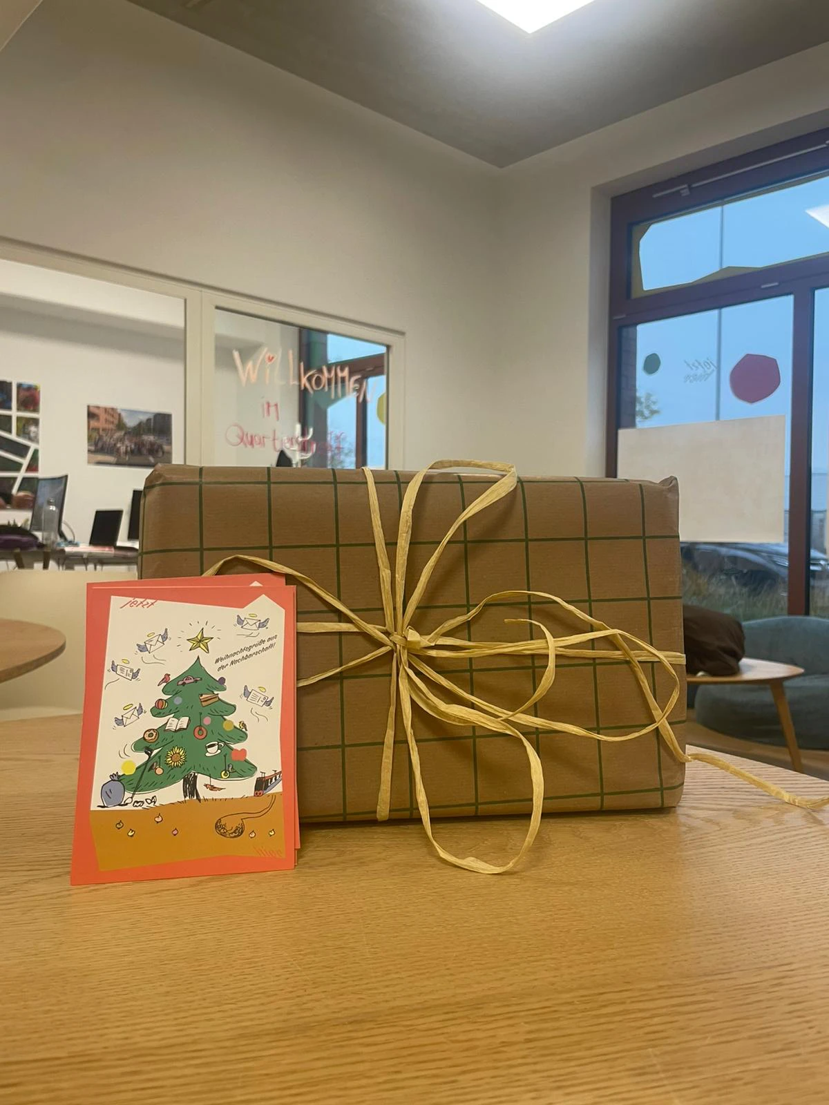 Zu sehen ist ein eingepacktes Geschenk mit einer Weihnachskarte auf einem Tisch. Im Hintergrund sind die Räumlichkeiten des Quartierstreff zu sehen.