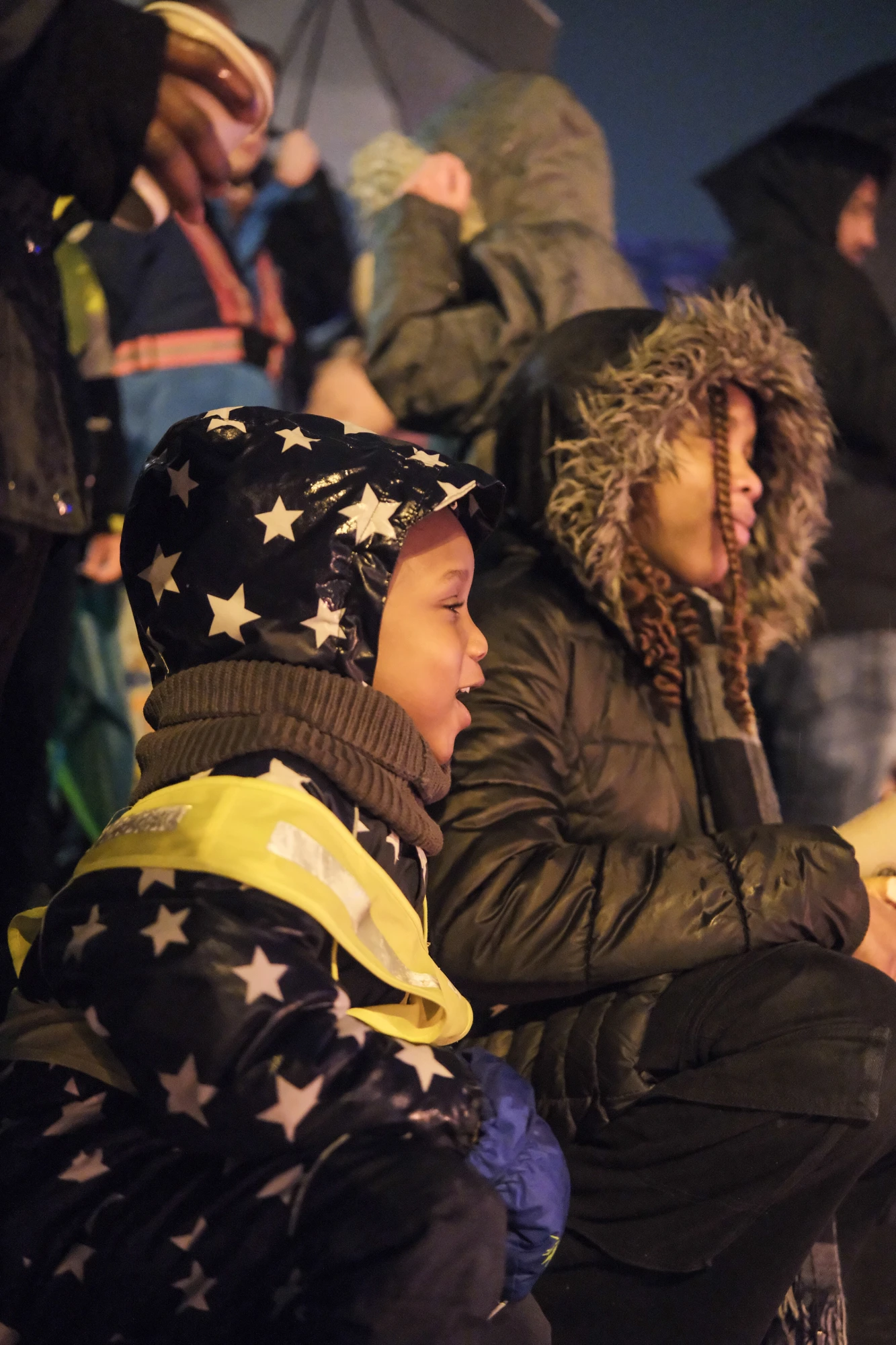 Zwei Kinder in Winterjacken sitzen nebeneinander, eines trägt eine Jacke mit Sternenmuster und eine gelbe Warnweste.