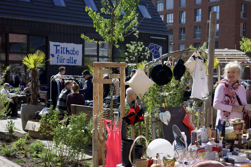 Marktstand mit verschiedenen Waren und Menschen im Hintergrund vor einem Gebäude mit einem Schild 'Teilhabe für alle'