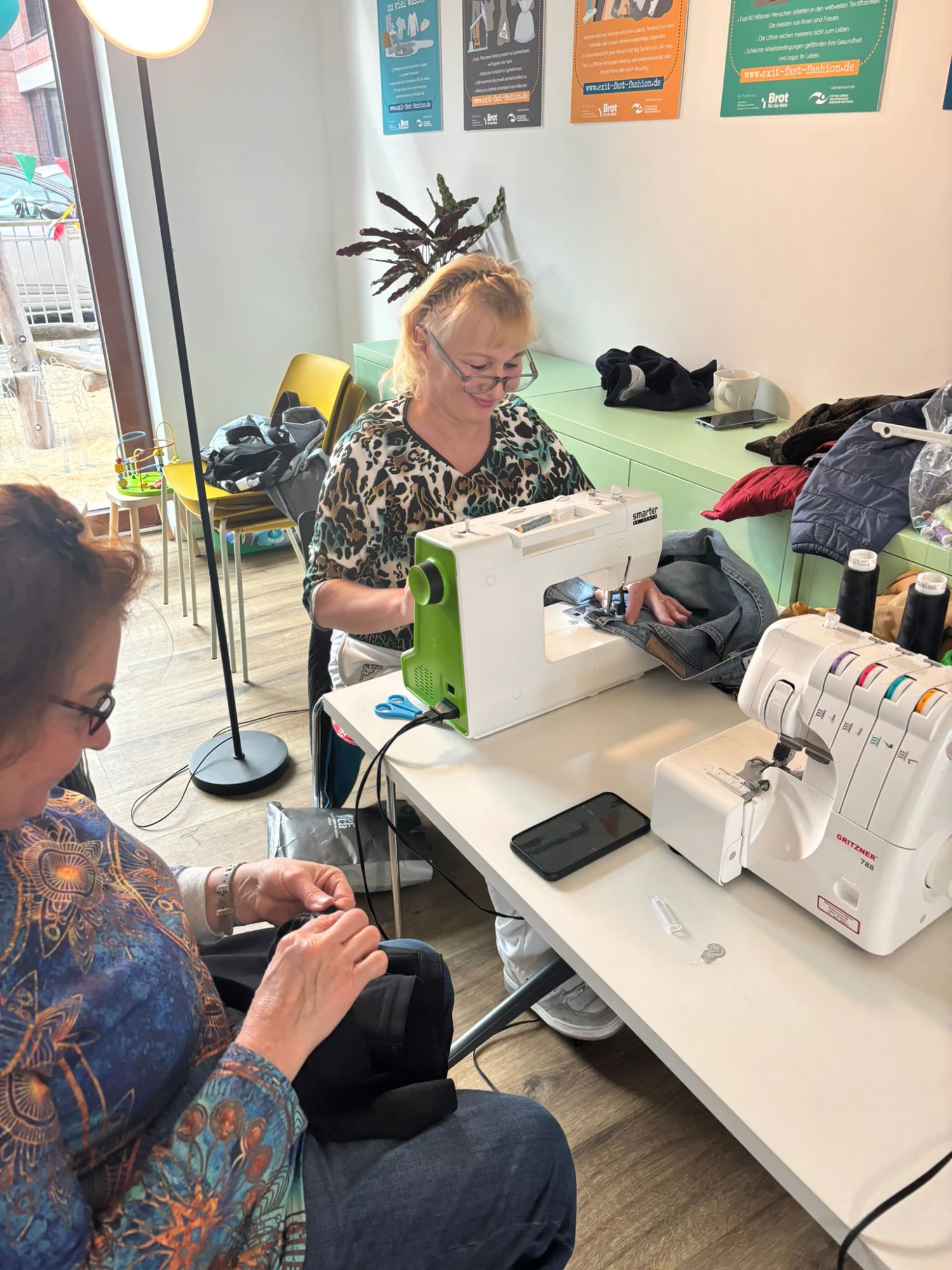Zwei Personen sitzen an einem Tisch mit zwei Nähmaschinen und nähen Kleidungsstücke in einem hellen Raum mit Poster an der Wand