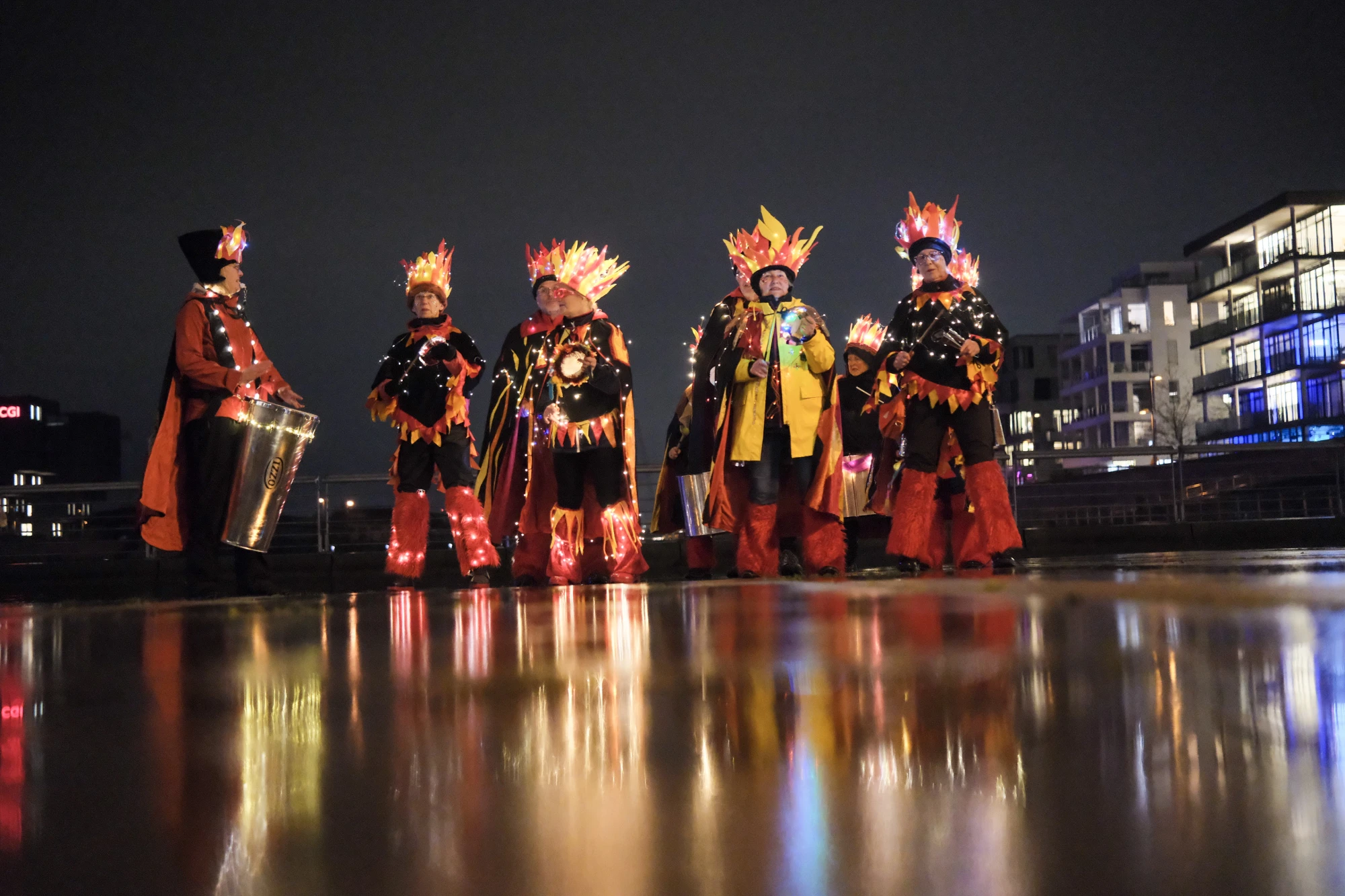 Gruppe von sechs Personen in leuchtenden Kostümen mit Flammenmotiven, eine Person spielt Trommel, nächtliche Stadtkulisse im Hintergrund.