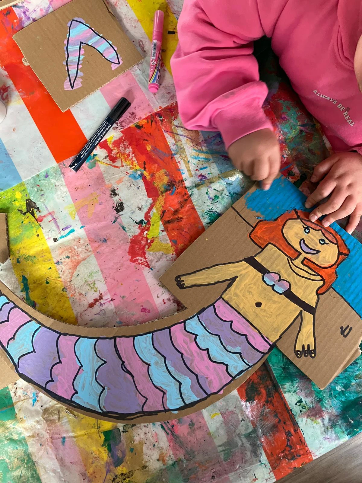 Eine bunt ausgemalte Meerjungfrau aus Pappe liegt auf einer bunten Tischdecke, die komplett mit Farbe voll ist. Ein Kind, dessen Hände zu sehen sind, malt gerade den HIntergrund blau an.