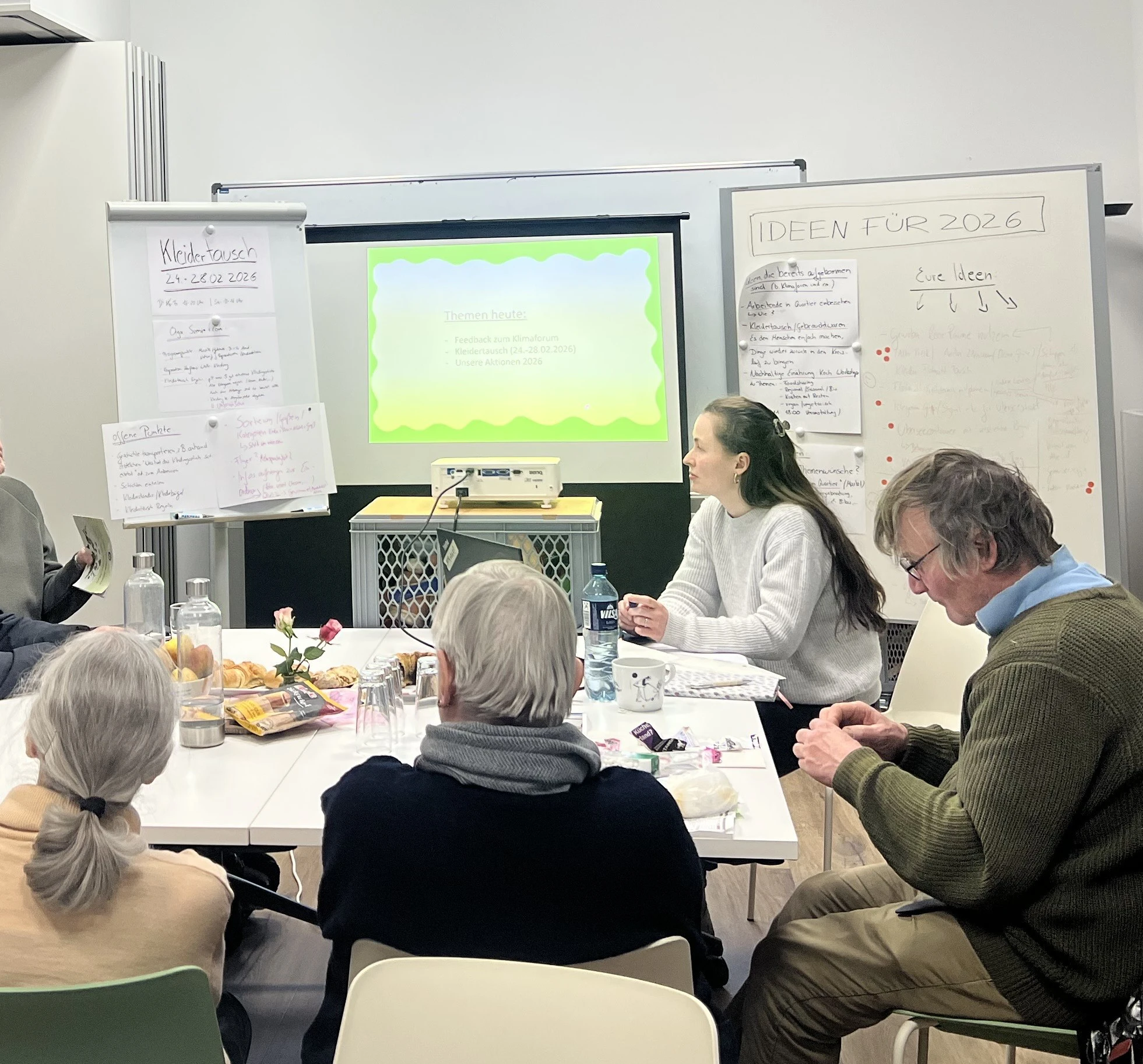 Gruppe von Menschen sitzt an einem Tisch mit Getr&auml;nken und Snacks vor Flipcharts und einem projizierten Bildschirm in einem Besprechungsraum