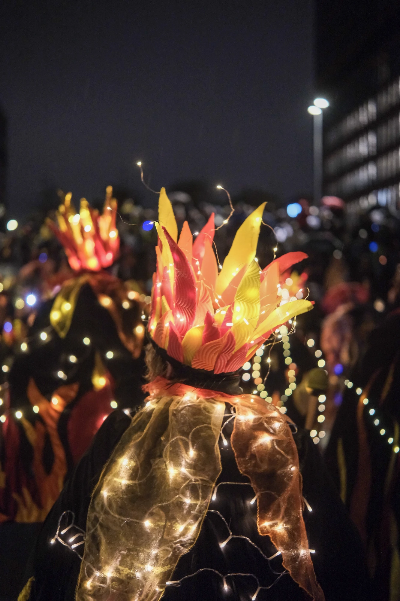Person mit leuchtender Kopfbedeckung in Flammenform und Lichterkette auf dem Rücken bei Nachtveranstaltung im Freien