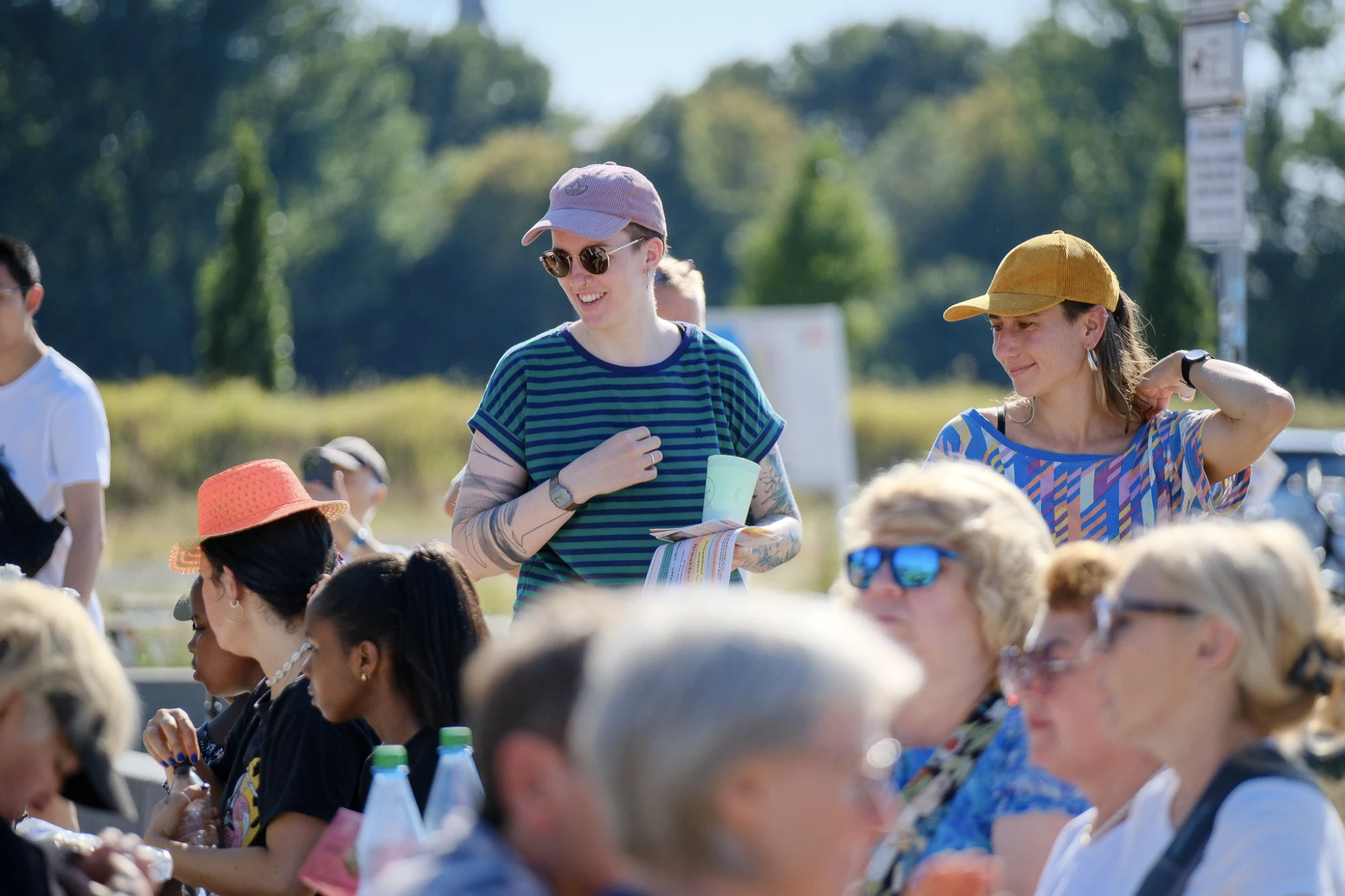 Im Vordergrund sitzen mehrere Menschen an einem langen Tisch im Freien. Sie tragen sommerliche Kleidung und unterhalten sich. Im Hintergrund stehen zwei Personen, die bunte Kappen und lässige Kleidung tragen. Eine Person hält einen Becher und lächelt. Die Szene spielt bei Tageslicht, im Hintergrund sind Bäume und ein Schild zu sehen.