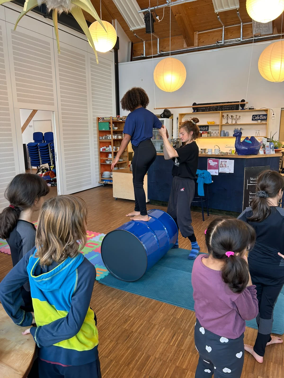 Kind balanciert auf einem blauen Fass, von einer erwachsenen Person gehalten, während mehrere Kinder zuschauen