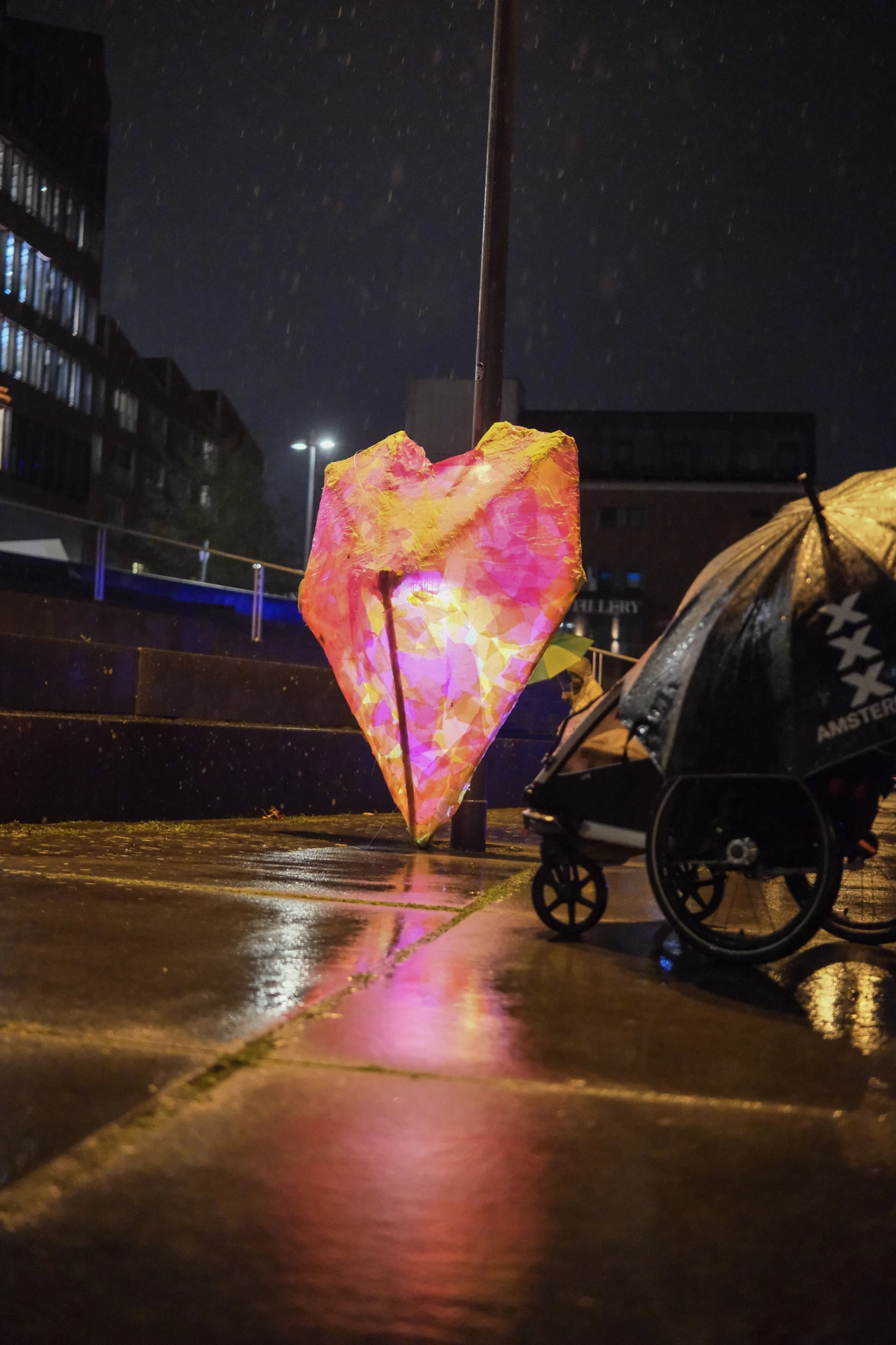 Leuchtende, herzförmige Laterne auf nassem Gehweg bei Nacht, daneben ein Kinderwagen mit Regenschutz und Amsterdam-Schriftzug.