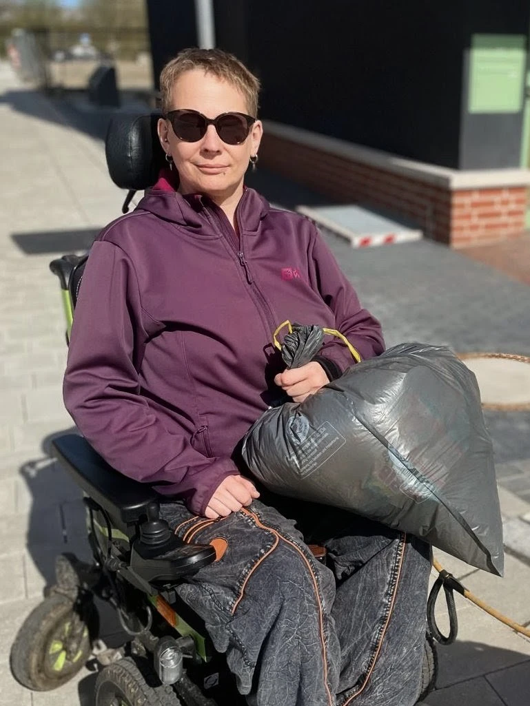 Zu sehen ist eine Person im Rollstuhl mit einem Müllsack in der Hand.