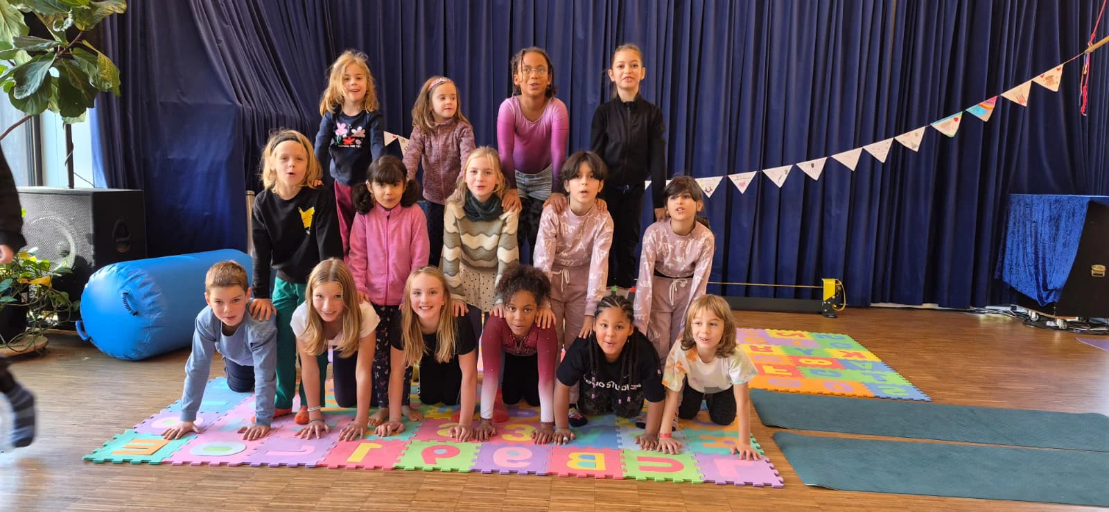Gruppe von Kindern in zwei Reihen auf bunten Spielmatten vor dunkelblauen Vorh&auml;ngen in einem Innenraum