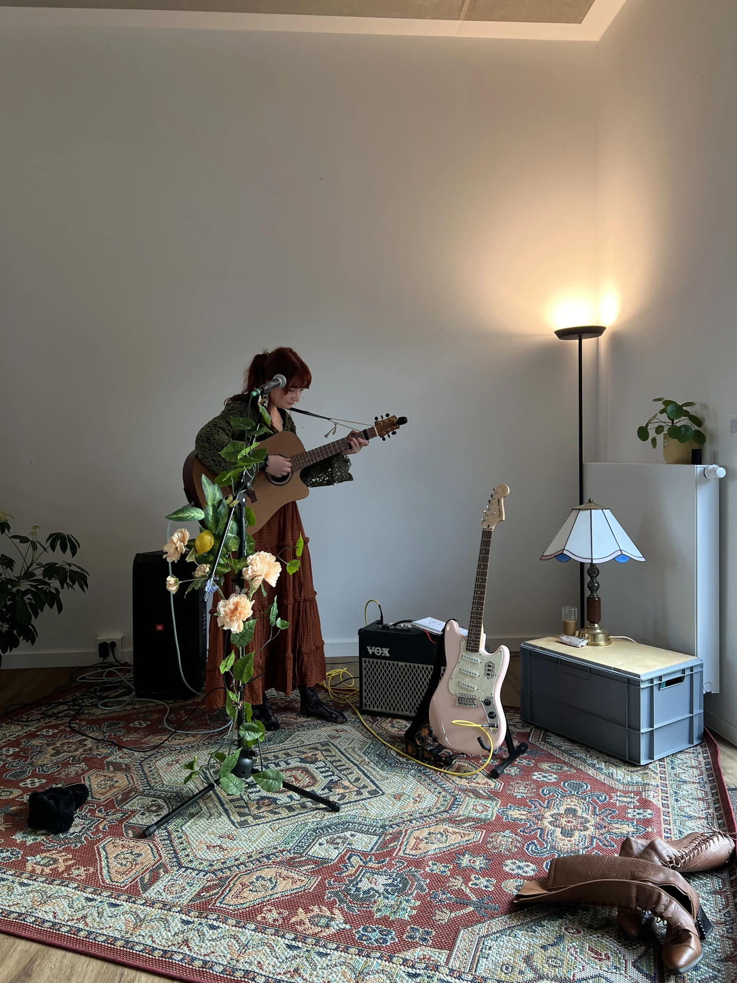 Person steht in einem Raum mit Akustikgitarre vor Mikrofonständer mit Blumen, daneben E-Gitarre auf Verstärker und Stehlampe auf Kiste