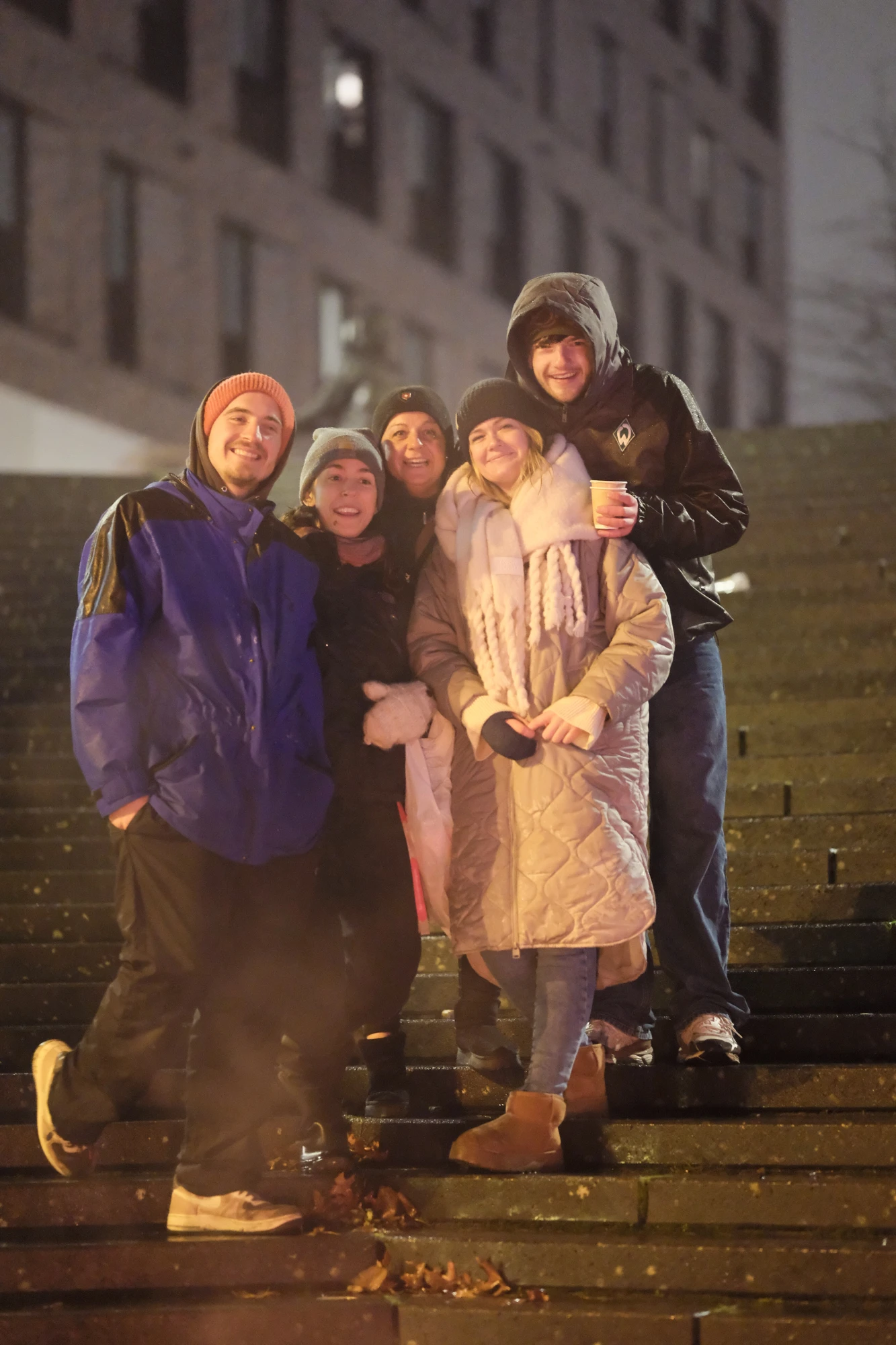Fünf Personen stehen auf einer Treppe bei Nacht, zwei tragen helle Mäntel, eine Person hält einen Becher.