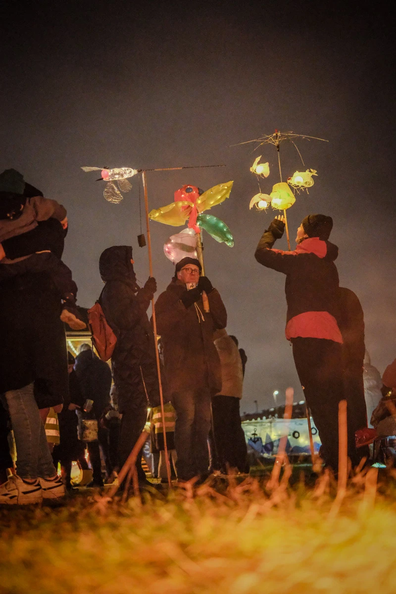 Auch einer Wiese stehen drei Menschen. Sie halten besondere Laternen an langen stäben. Eine sieht aus wie ein Insekt, eine andere wie eine  Blume. Die dritte besteht aus kleineren leuchtenden Lampen, die an einem kaputten Schirm befestigt sind.