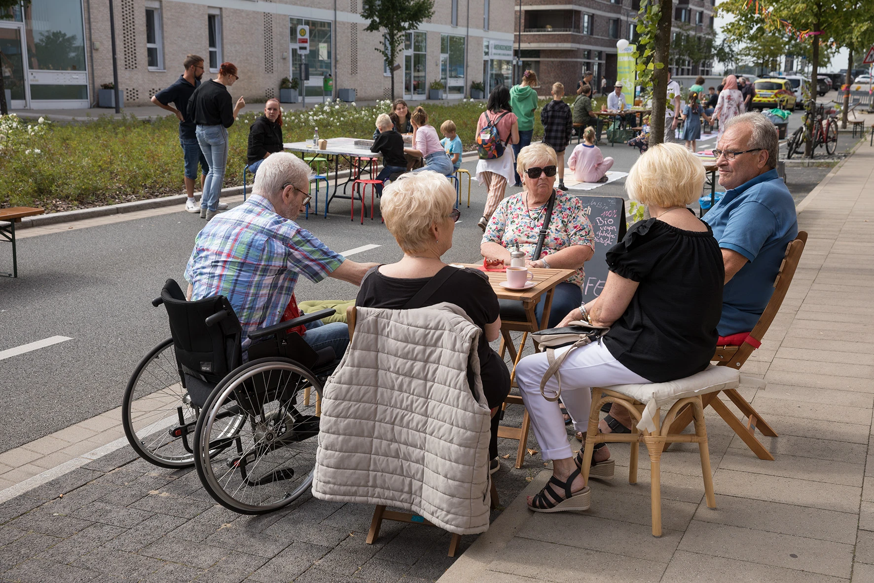 Gruppe von Menschen sitzt an Tischen auf einem Bürgersteig neben einer Straße in einem urbanen Gebiet