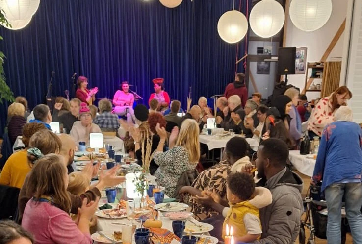 Viele Menschen sitzen am gedeckten Tisch, applaudieren und schauen Richtung B&uuml;hne. Hier sitzen drei Frauen.