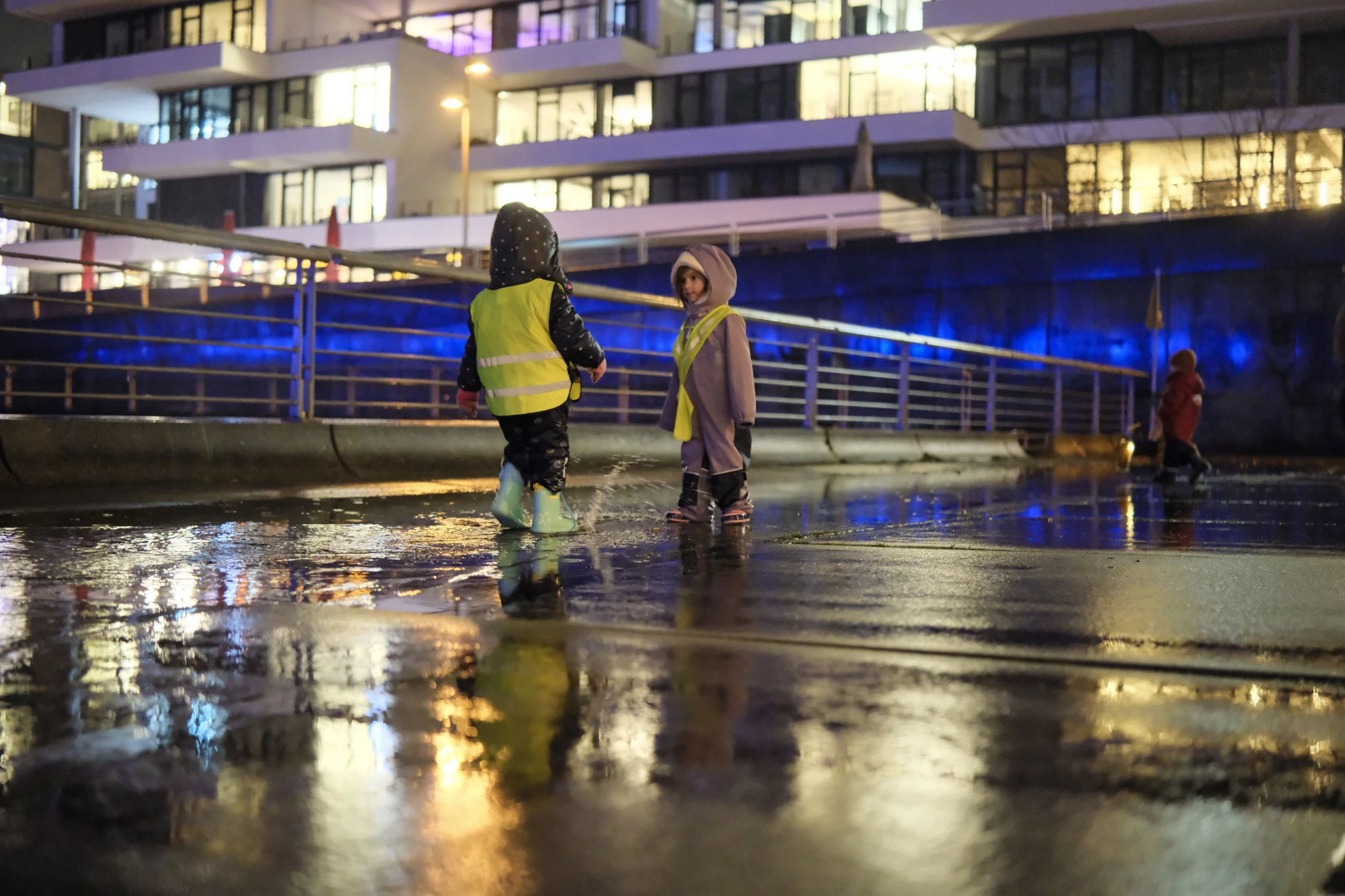 Kinder mit Kapuzenjacke und gelber Warnweste läuft auf nasser Straße mit Reflexionen bei Nacht.