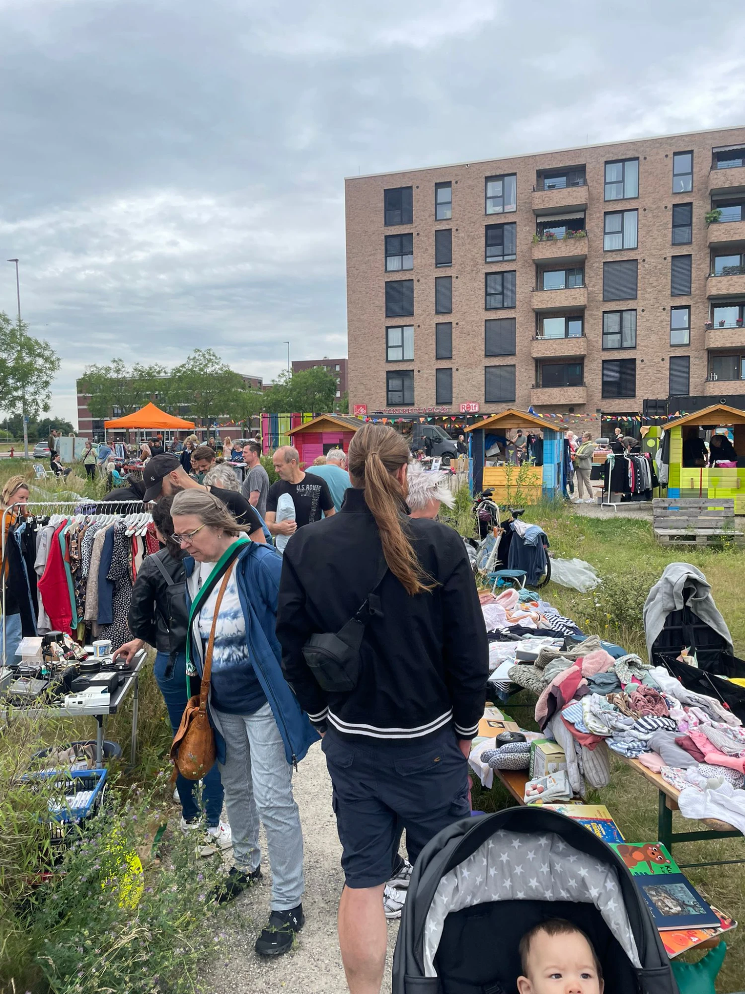 Im Vordergrund stehen mehrere Menschen auf einem schmalen Kiesweg, der zwischen dicht bewachsenen Gr&uuml;nfl&auml;chen verl&auml;uft. Links und rechts des Weges sind Tische und St&auml;nder mit Kleidung, Spielzeug und Haushaltswaren aufgebaut. Im Hintergrund sind bunte Marktst&auml;nde und ein mehrst&ouml;ckiges Backsteingeb&auml;ude mit Balkonen zu sehen. Der Himmel ist bew&ouml;lkt.