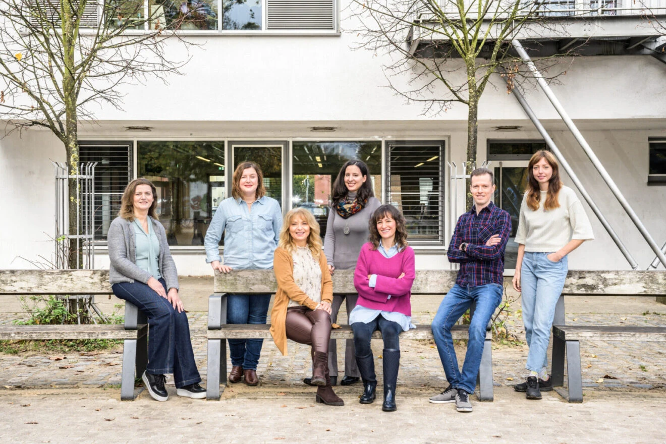 Sieben Personen stehen vor dem Quartierszentrum Morgenland in Gröpelingen. Sie stehen versammelt um Bänke herum und lachen in die Kamera. Es ist das Team der Bildungsberatung in Gröpelingen.