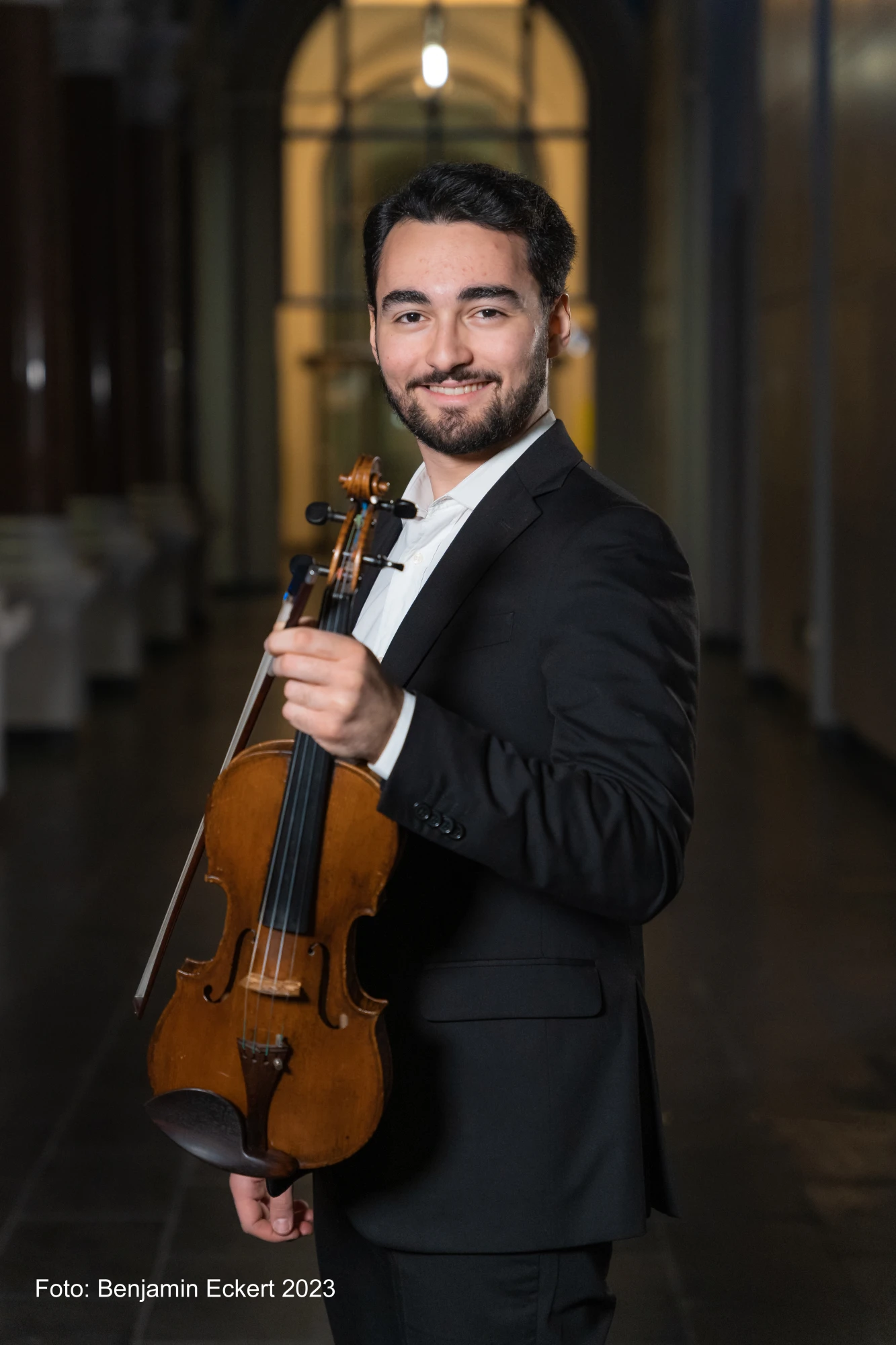 Ein junger Mann im Anzug steht in einem beleuchteten Innenraum und hält eine Geige in der linken Hand. Er lächelt in die Kamera.