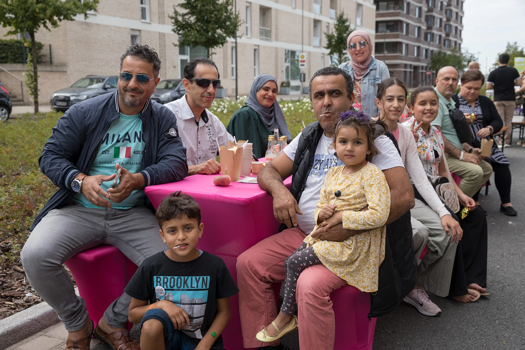 Mehrere Menschen sitzen und stehen an einem pinkfarbenen Tisch im Freien auf einer asphaltierten Fläche. Im Hintergrund sind moderne mehrstöckige Wohngebäude und einige Bäume zu sehen. Auf dem Tisch stehen Pappbecher und Essensverpackungen. Die Personen tragen legere Kleidung, darunter Jacken, T-Shirts und ein gemustertes Kleid. Ein Kind sitzt auf dem Schoß eines Erwachsenen, ein weiteres Kind sitzt auf dem Boden vor dem Tisch. Die Szene wirkt wie ein nachbarschaftliches Treffen oder eine Veranstaltung im Freien.