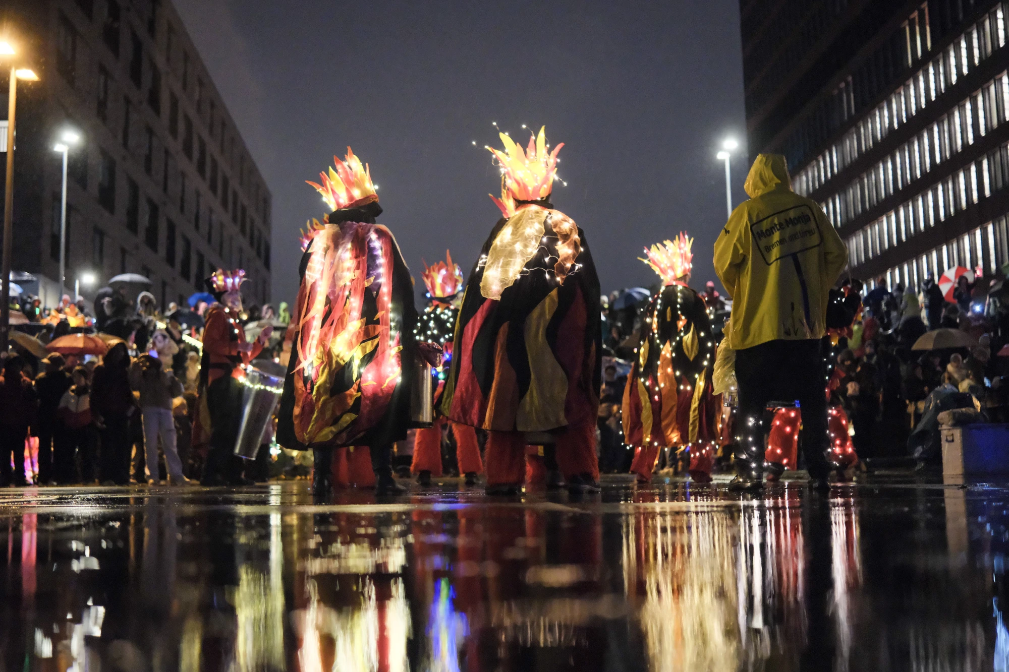 Mehrere Personen in leuchtenden Kostümen mit Flammenmuster und Kronen aus Licht stehen auf nassem Pflaster bei Nacht, im Hintergrund Zuschauer und Straßenlaternen.