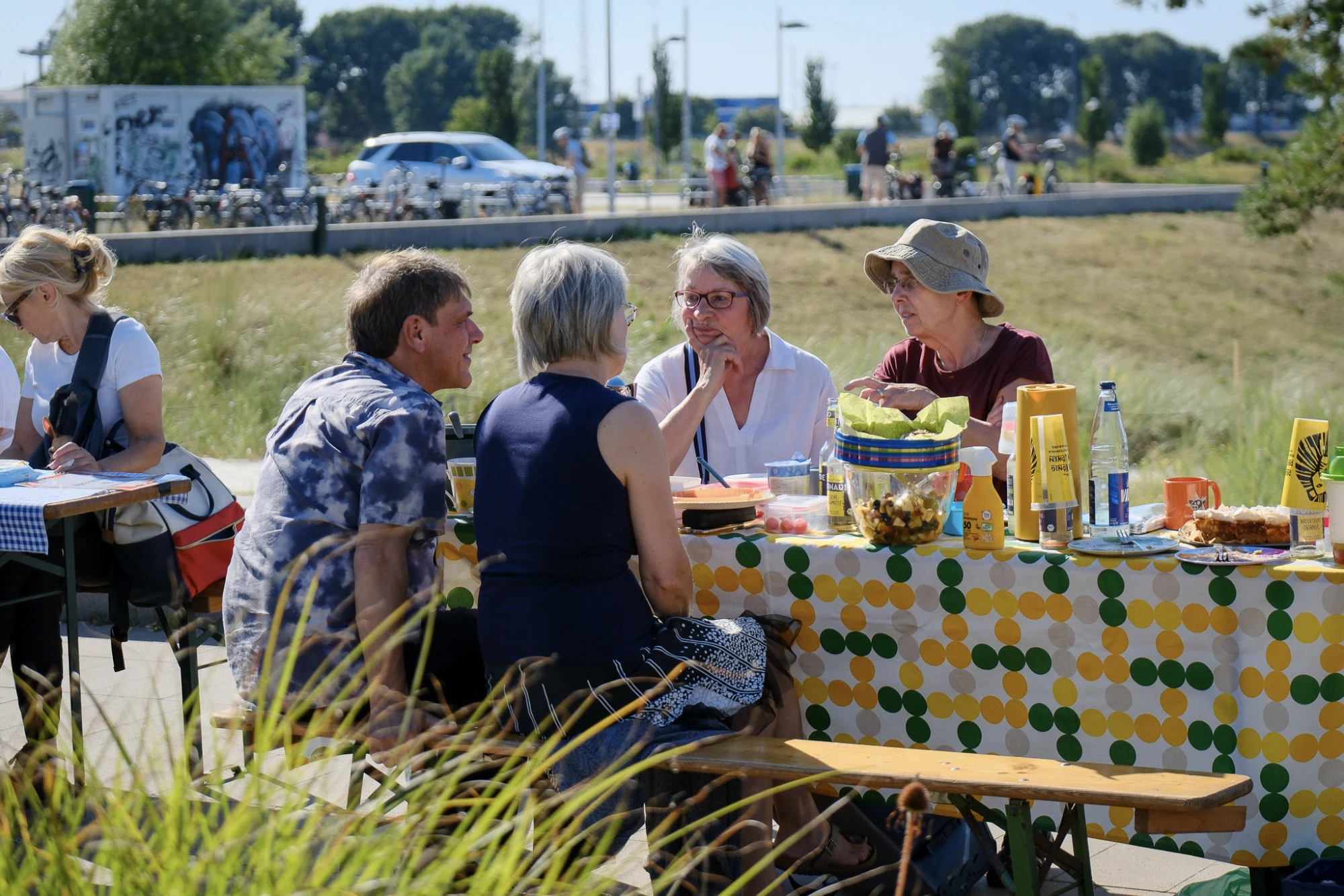 Im Vordergrund sitzen mehrere Menschen an einem langen Holztisch im Freien. Der Tisch ist mit einer bunten Tischdecke mit gelben und grünen Kreisen bedeckt und mit Schalen, Flaschen und Servietten bestückt. Die Personen unterhalten sich und wirken entspannt. Im Hintergrund sind eine Wiese, einige Bäume, ein Graffiti-Wand und parkende Fahrräder sowie Autos zu sehen. Die Szene spielt bei sonnigem Wetter.