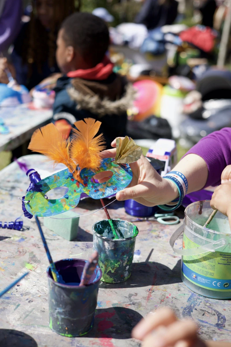 Hand hält bunte Maske mit orangen Federn über bemalten Bechern und Pinseln auf einem Tisch im Freien
