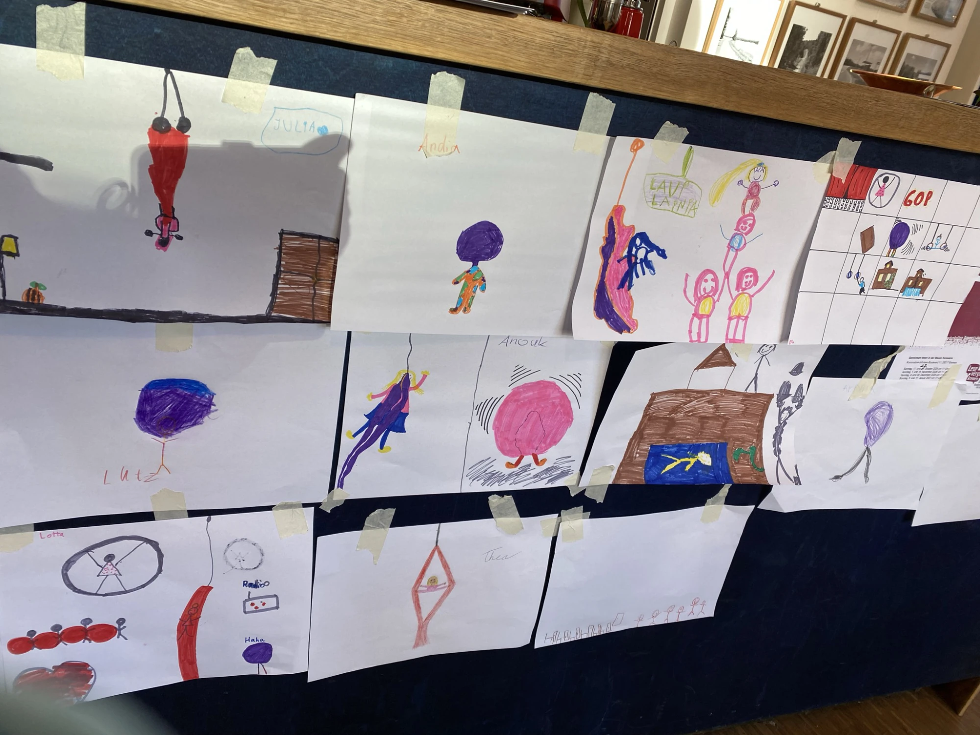Mehrere Kinderzeichnungen mit Figuren, Häusern und abstrakten Formen an einer Wand befestigt.