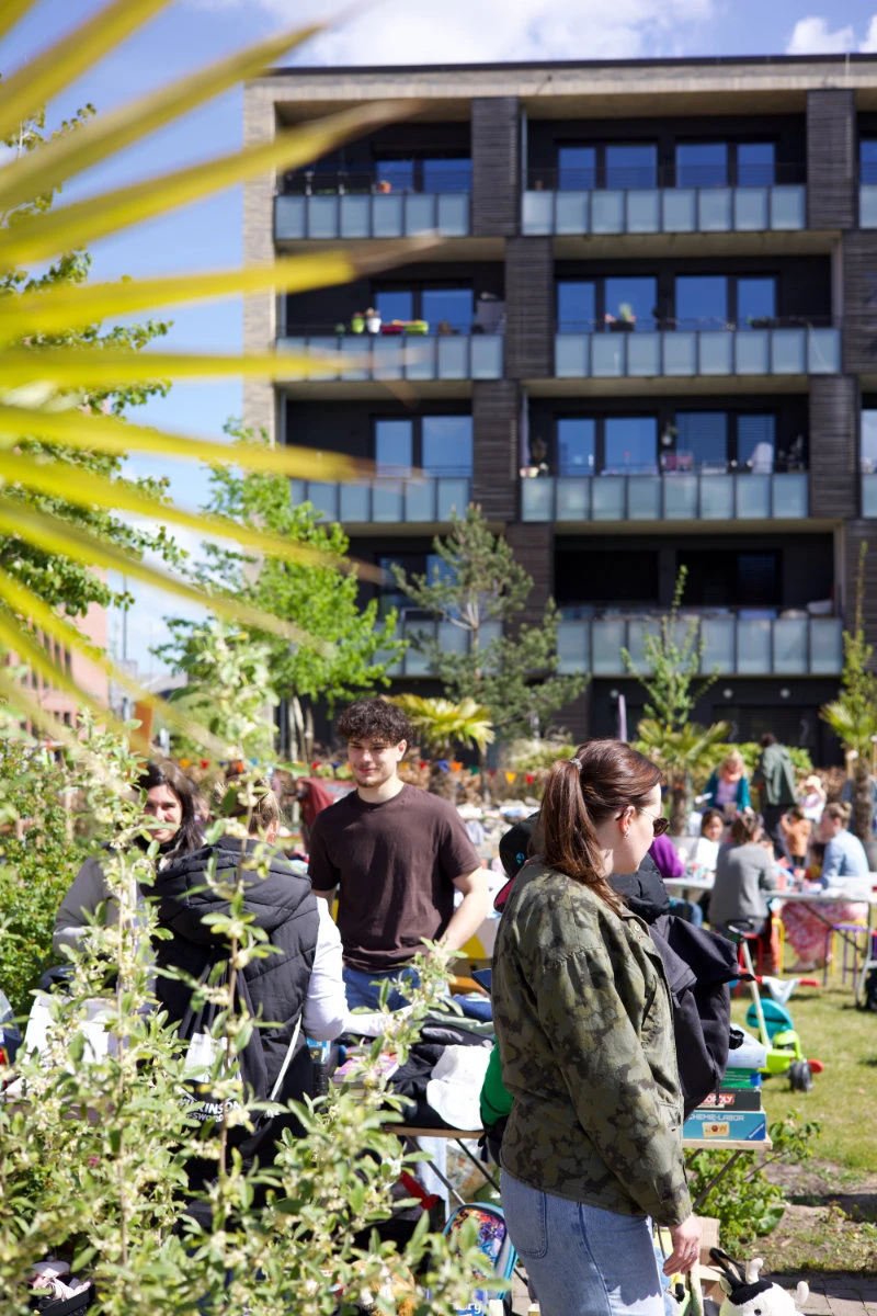 Menschen sitzen und stehen auf einer Wiese vor einem modernen Wohngebäude mit Balkonen an einem sonnigen Tag