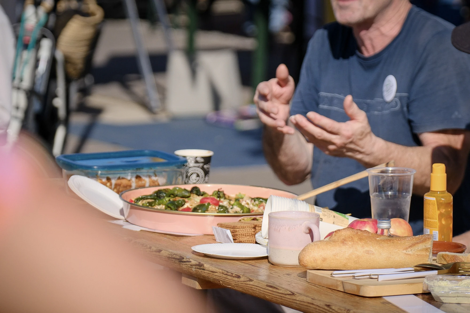 Mann mit weißen Haaren und Sonnenbrille sitzt im Rollstuhl und gestikuliert vor einem Tisch mit Schüssel voller Salat