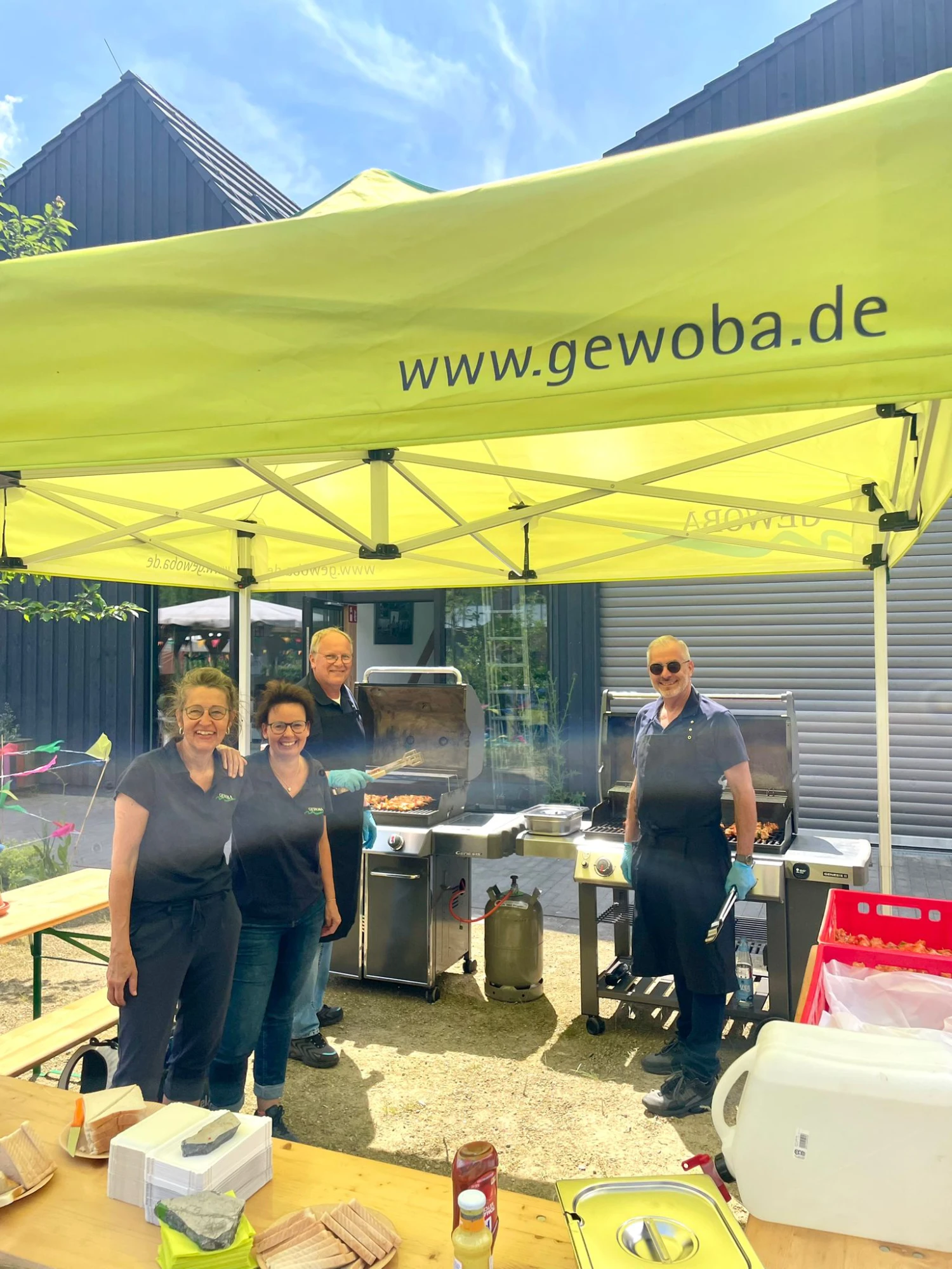 Drei Personen stehen unter einem gelben Pavillon mit der Aufschrift 'www.gewoba.de' vor einem Grill und Tischen mit Grillzubehör.