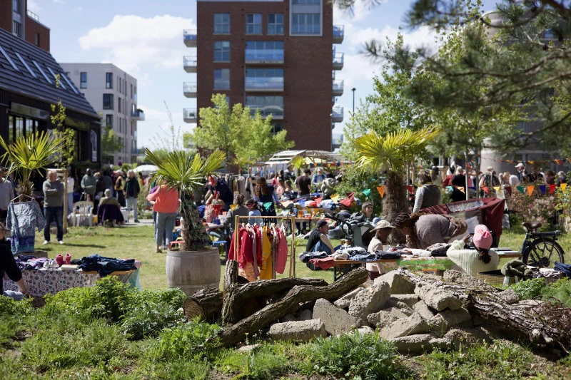28.	Menschen verschiedener Altersgruppen stehen und gehen auf einem sonnigen Platz vor einem modernen Wohngeb&auml;ude mit Balkonen und B&auml;umen