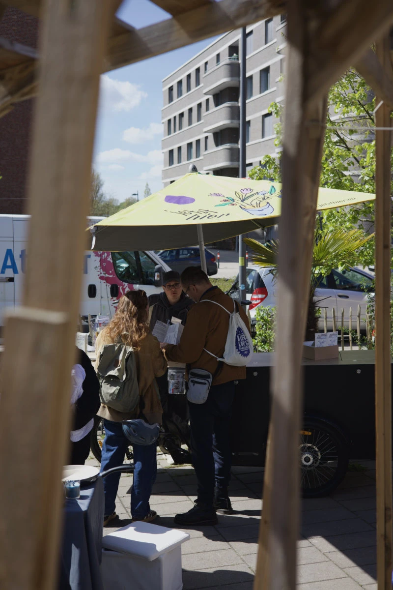 Gruppe von Menschen steht unter einem gelben Sonnenschirm auf einem Bürgersteig vor einem modernen Gebäude