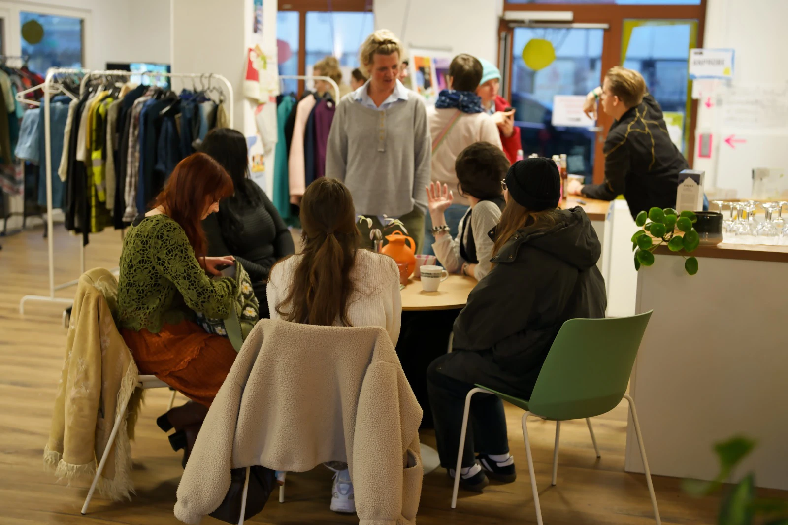 Gruppe von Menschen sitzt und steht in einem hellen Raum mit Garderobenständern und Pflanzen, einige unterhalten sich am Tisch