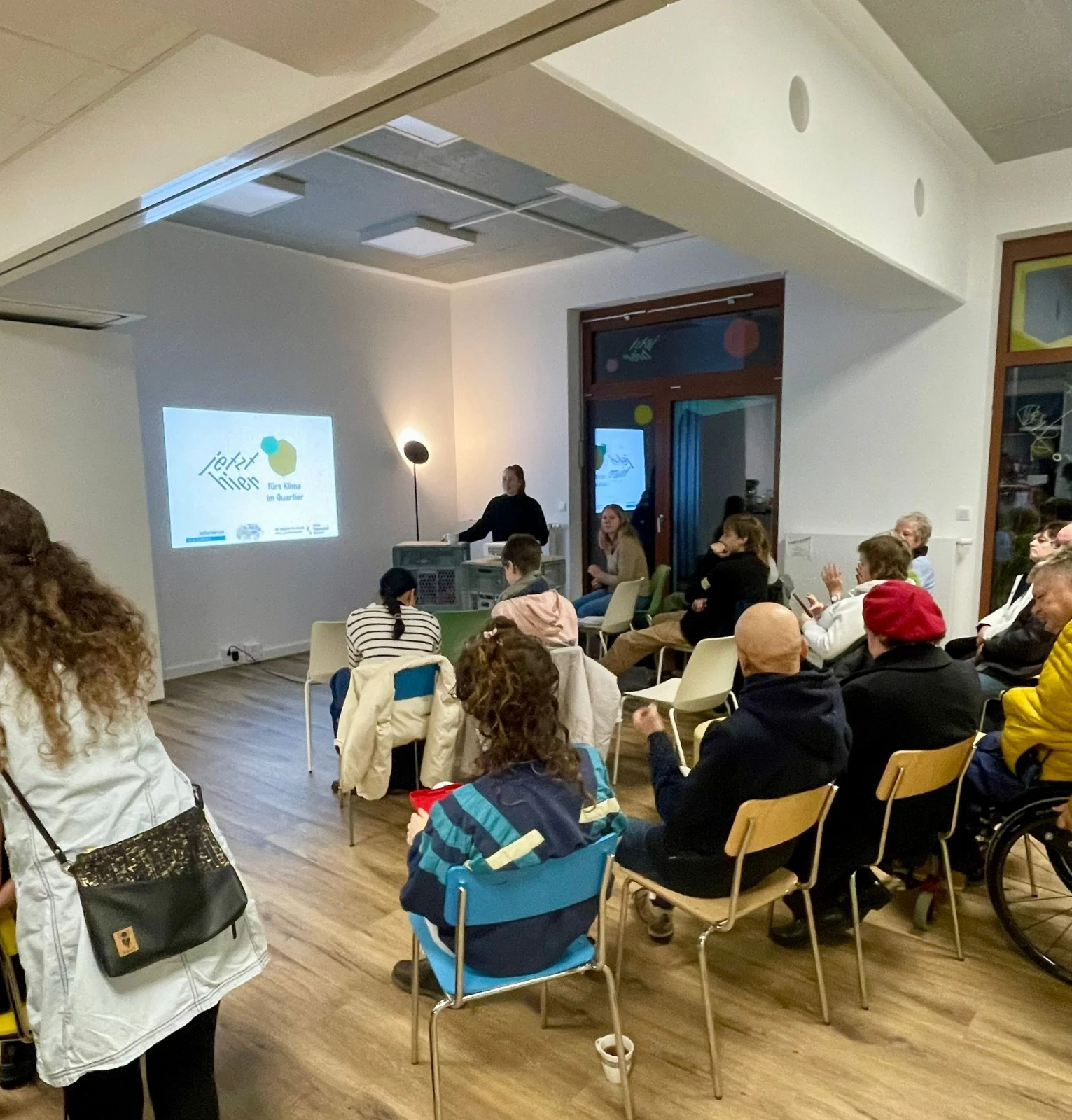 Mehrere Personen sitzen in einem Raum mit Holzfu&szlig;boden und wei&szlig;en W&auml;nden und h&ouml;ren einer Person zu, die vor einem Projektor steht, der eine Pr&auml;sentation an die Wand wirft