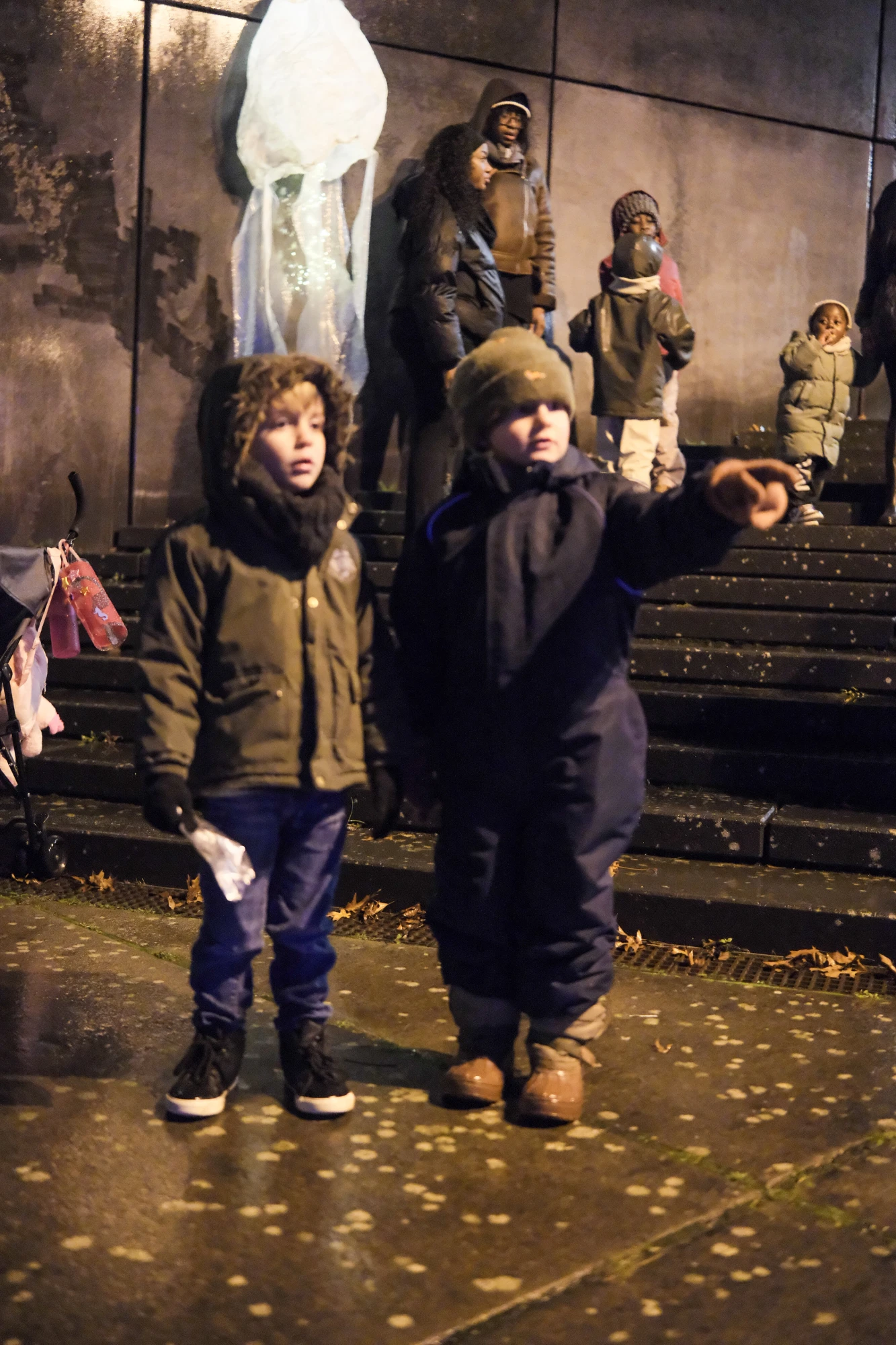 Zwei Kinder in Winterkleidung stehen auf nassem Boden, eines zeigt mit ausgestrecktem Arm nach rechts, im Hintergrund weitere Personen auf Treppen vor einer Wand mit großem, leuchtendem Laternenbild.