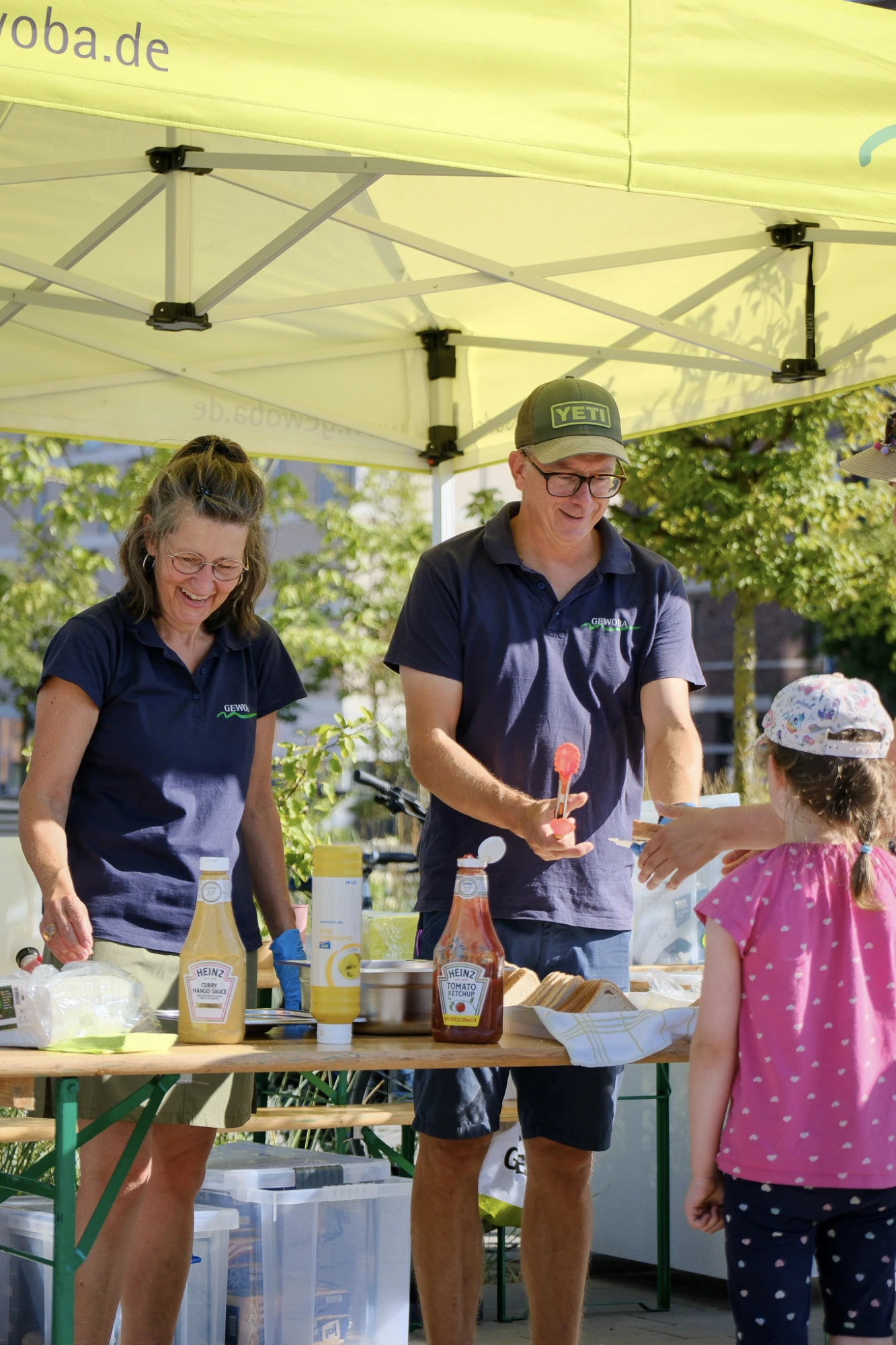 Im Vordergrund steht ein Mädchen mit hellem T-Shirt und gemusterter Kappe vor einem Holztisch. Dahinter stehen zwei Erwachsene unter einem gelben Pavillon, beide tragen dunkelblaue Poloshirts und lächeln. Auf dem Tisch befinden sich mehrere Flaschen mit Saucen, Becher und weitere Lebensmittel. Die Szene spielt im Freien bei sonnigem Wetter.