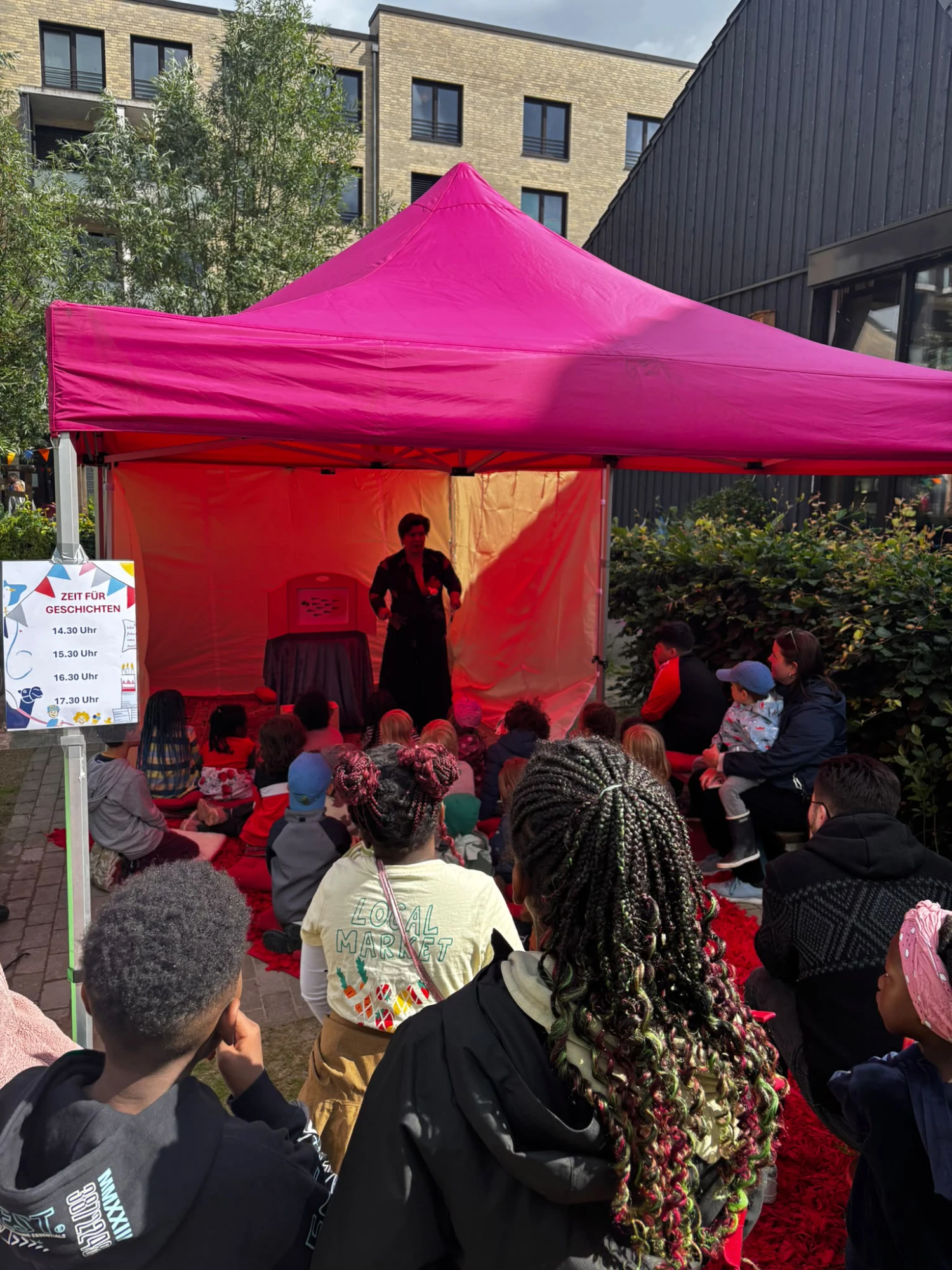 Kinder sitzen vor einem roten Pavillon, in dem eine Person steht und Geschichten erzählt.