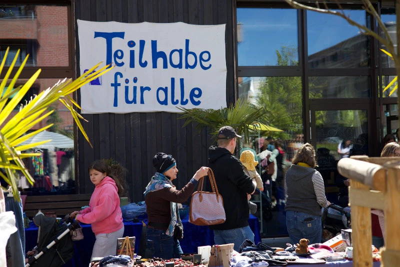 Menschen stehen an einem Marktstand vor einem Gebäude mit einem Banner, auf dem 'Teilhabe für alle' steht