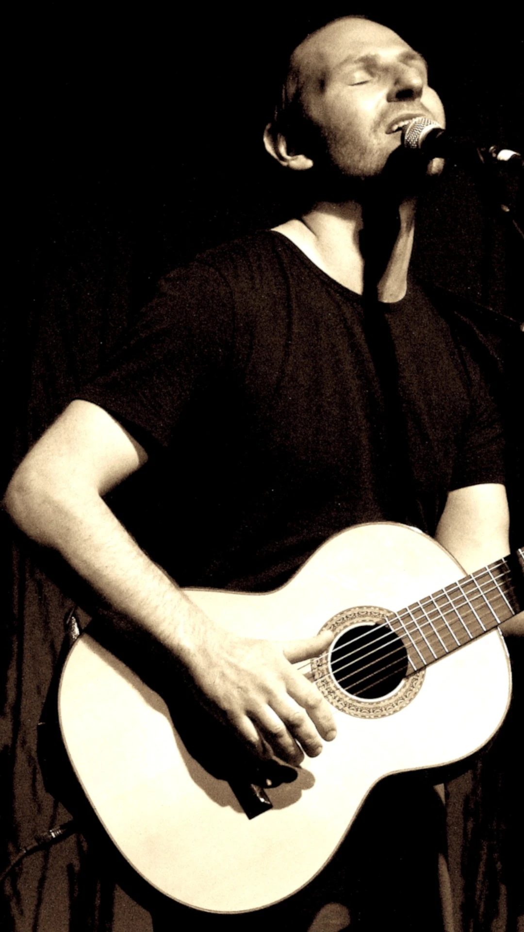 Person in schwarzem T-Shirt spielt akustische Gitarre vor dunklem Hintergrund