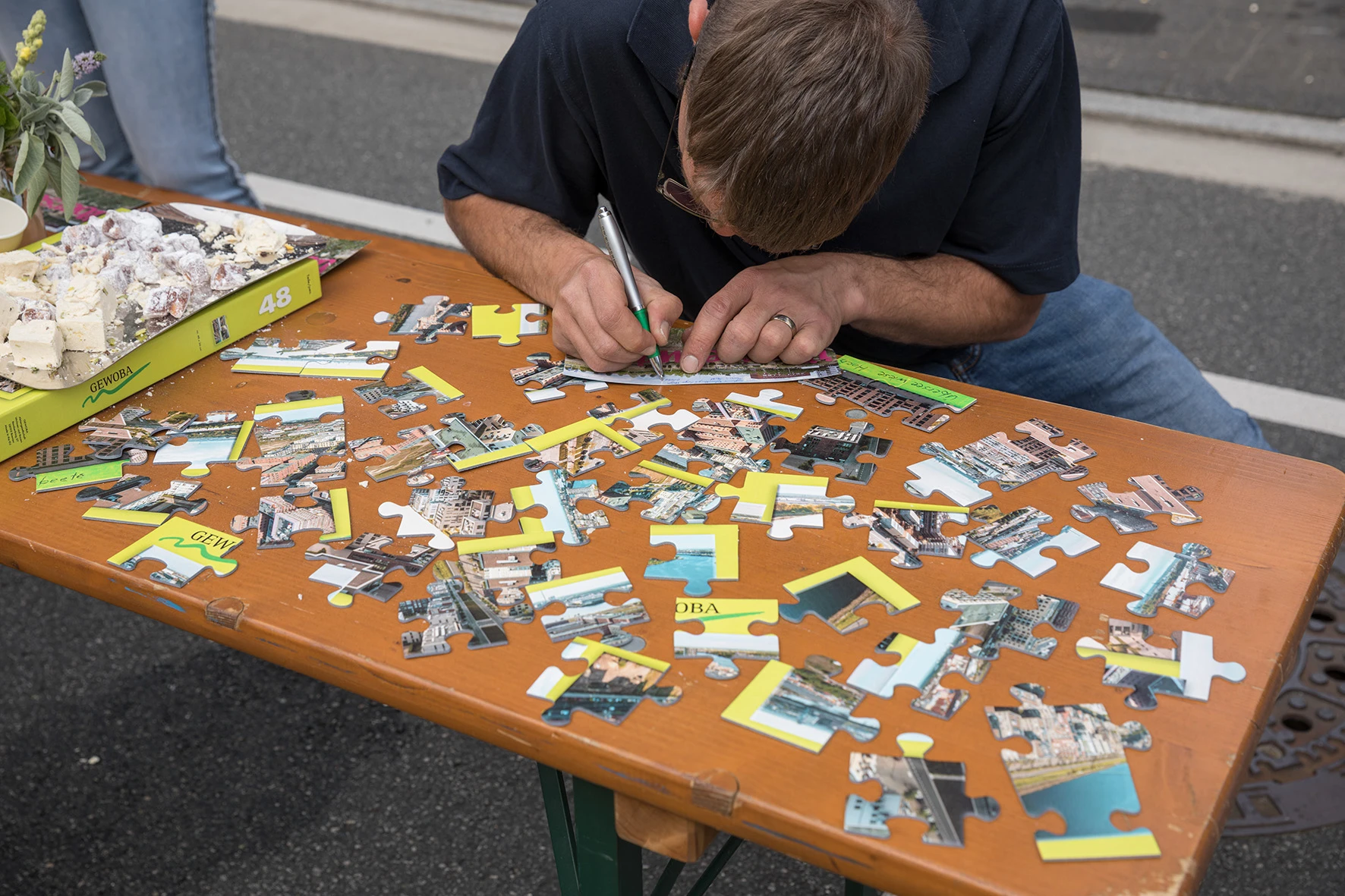 Ein Mann mit kurz geschnittenem Haar und Brille beugt sich über einen Holztisch und beschriftet mit einem Stift ein großes Puzzleteil. Auf dem Tisch liegen zahlreiche weitere, übergroße Puzzleteile mit Fotomotiven und gelben Flächen verteilt. Am linken Bildrand steht eine offene Puzzle-Schachtel mit der Aufschrift '48', daneben ein Tablett mit Gebäckstücken und eine kleine Blumenvase.