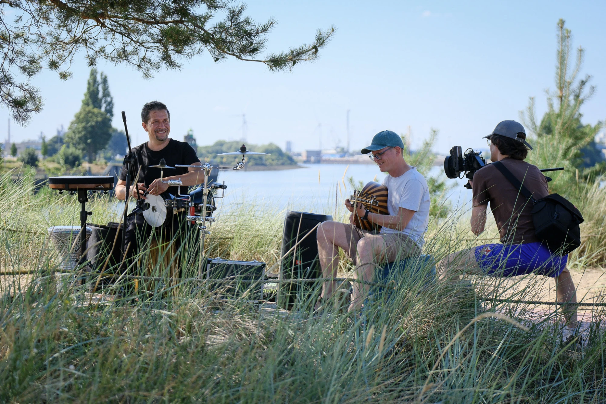 Zwei Personen sitzen im hohen Gras am Ufer eines Flusses. Eine Person spielt ein Schlaginstrument, die andere hält ein weiteres Musikinstrument in den Händen. Beide sind leger gekleidet. Im Vordergrund steht eine weitere Person mit Kameraausrüstung und filmt die Szene. Im Hintergrund sind Wasser, Bäume und einige Bauwerke zu sehen. Die Szene wirkt sommerlich und entspannt.