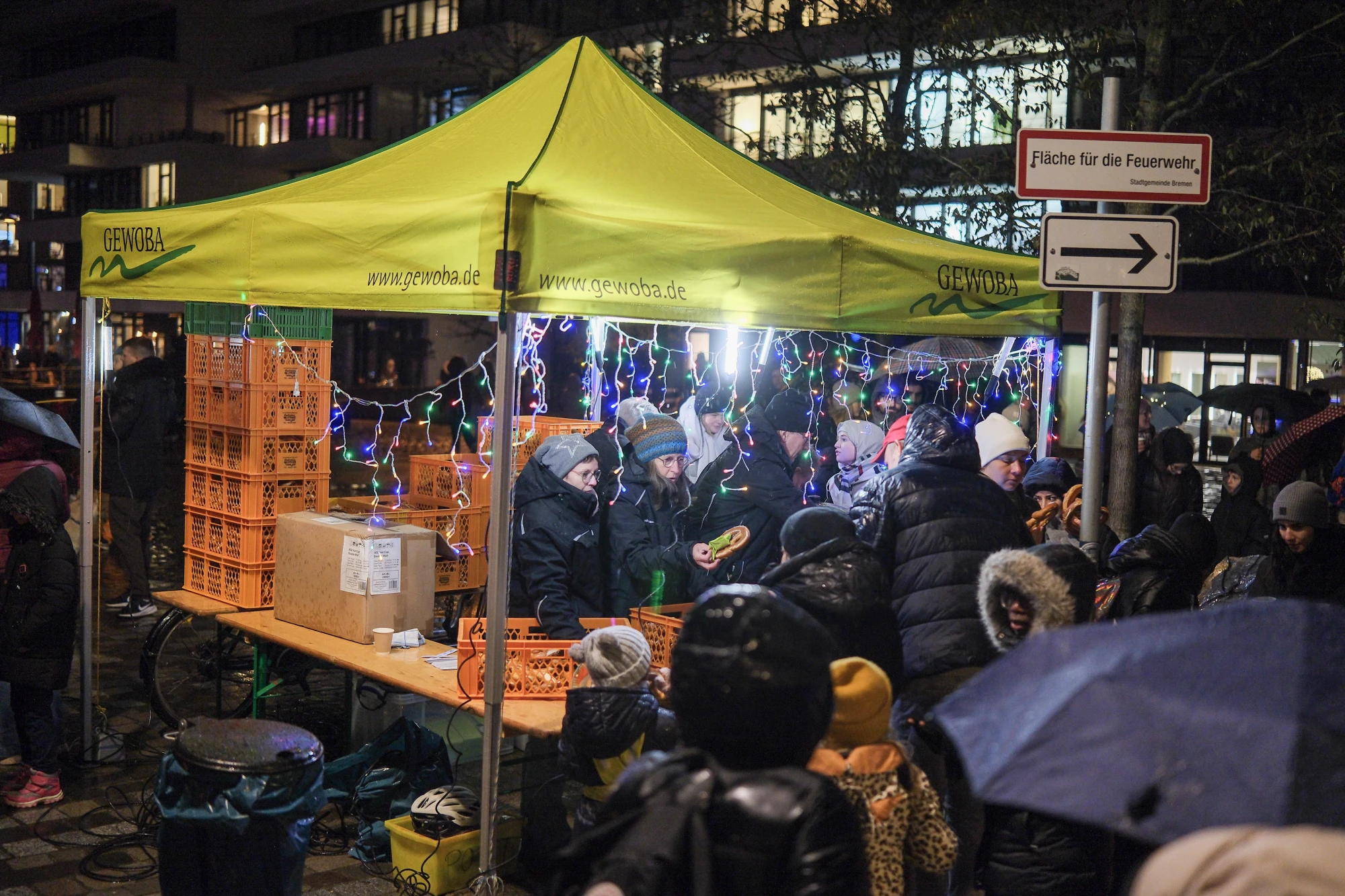 Gelbes Zelt mit Aufschrift 'GEWOBA' und 'www.gewoba.de', darunter Menschen mit Winterkleidung und Weihnachtsbeleuchtung im Hintergrund.