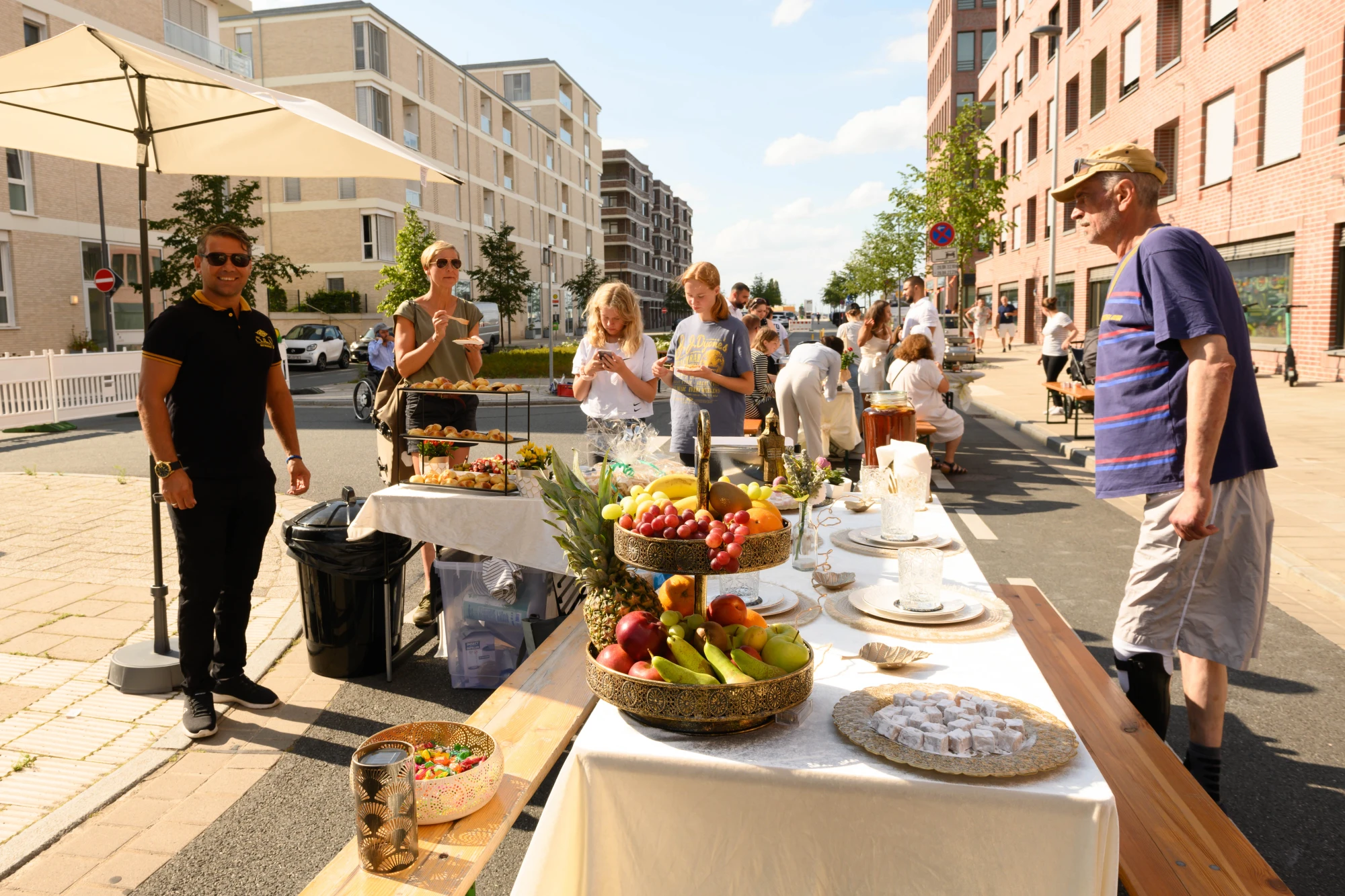 Straßenszene mit mehreren Menschen an einem langen Tisch mit Obst, Gebäck und Getränken unter Sonnenschirmen in einem urbanen Viertel