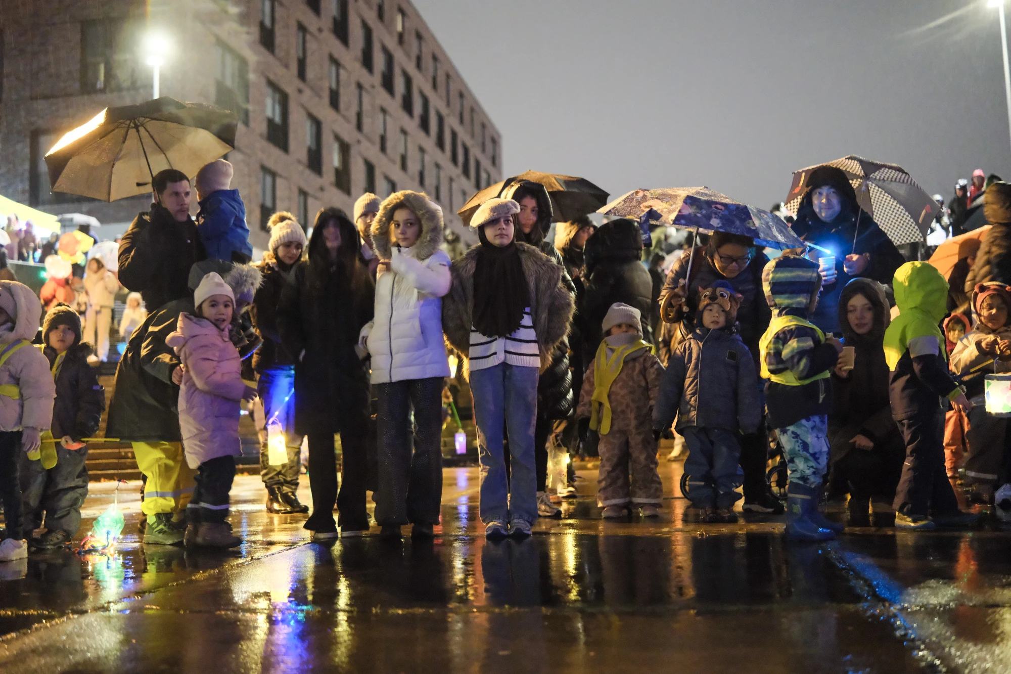 Gruppe von Kindern und Erwachsenen mit Regenschirmen und Laternen bei Nacht auf nassem Boden vor beleuchtetem Gebäude.