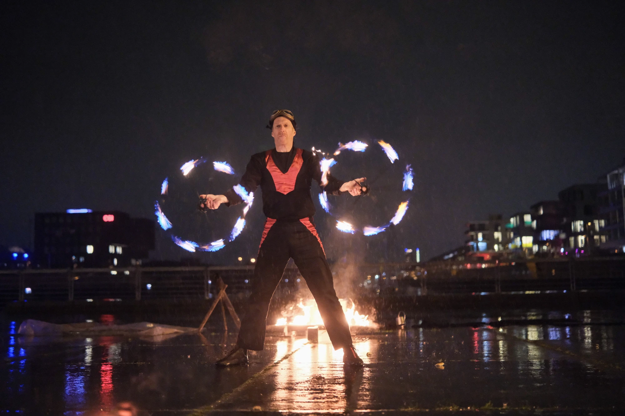 Person in dunkler Kleidung mit reflektierenden Streifen hält brennende Laterne über nassem Boden bei Nacht.